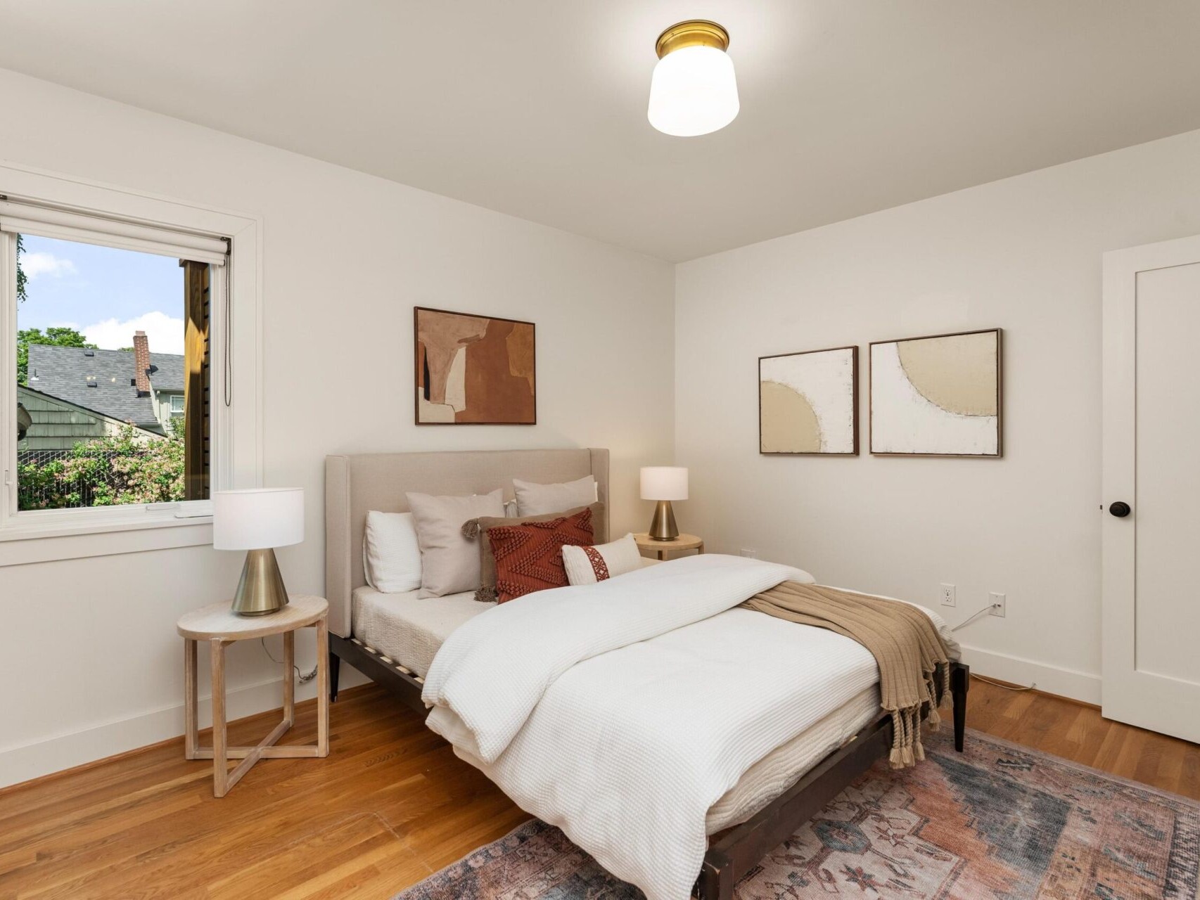 A cozy bedroom with a neatly made bed featuring white linens and brown accent pillows. Two abstract paintings hang on the white walls. A window allows natural light in, and two bedside lamps provide warm lighting. A soft rug covers the wooden floor.