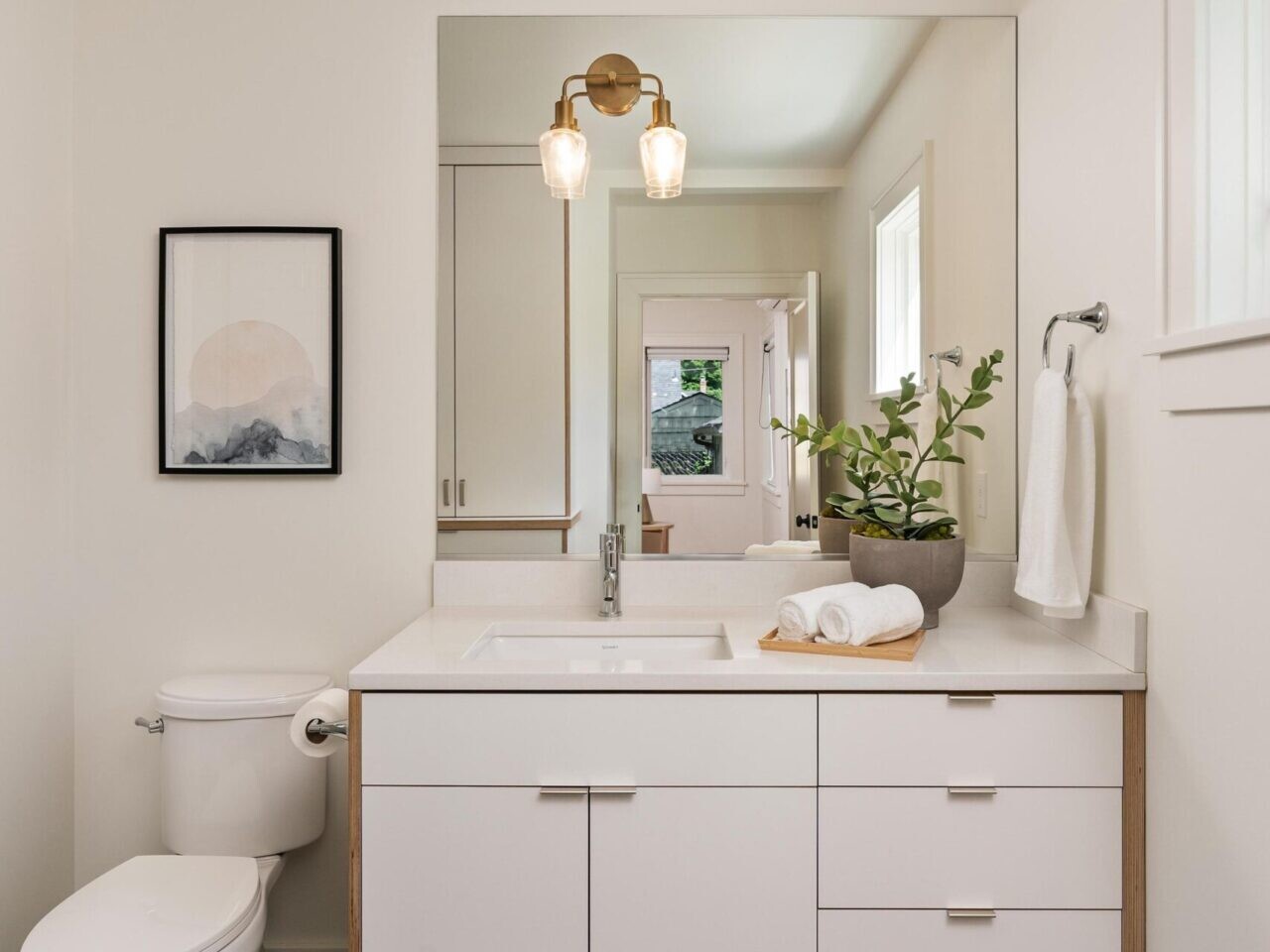A modern bathroom features a white vanity with a large mirror, under a two-light fixture. A toilet sits to the left, and a window to the right. Decor includes a potted plant, neatly folded towels, and a small framed artwork. The floor has hexagonal tiles.