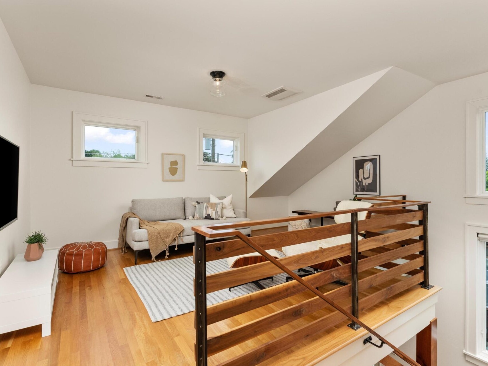 A bright attic living space with wooden flooring. It features a cozy gray sofa with cushions, a small white table, and a brown pouf. A flat-screen TV is mounted on the wall, and framed art decorates the walls. The room has several windows.