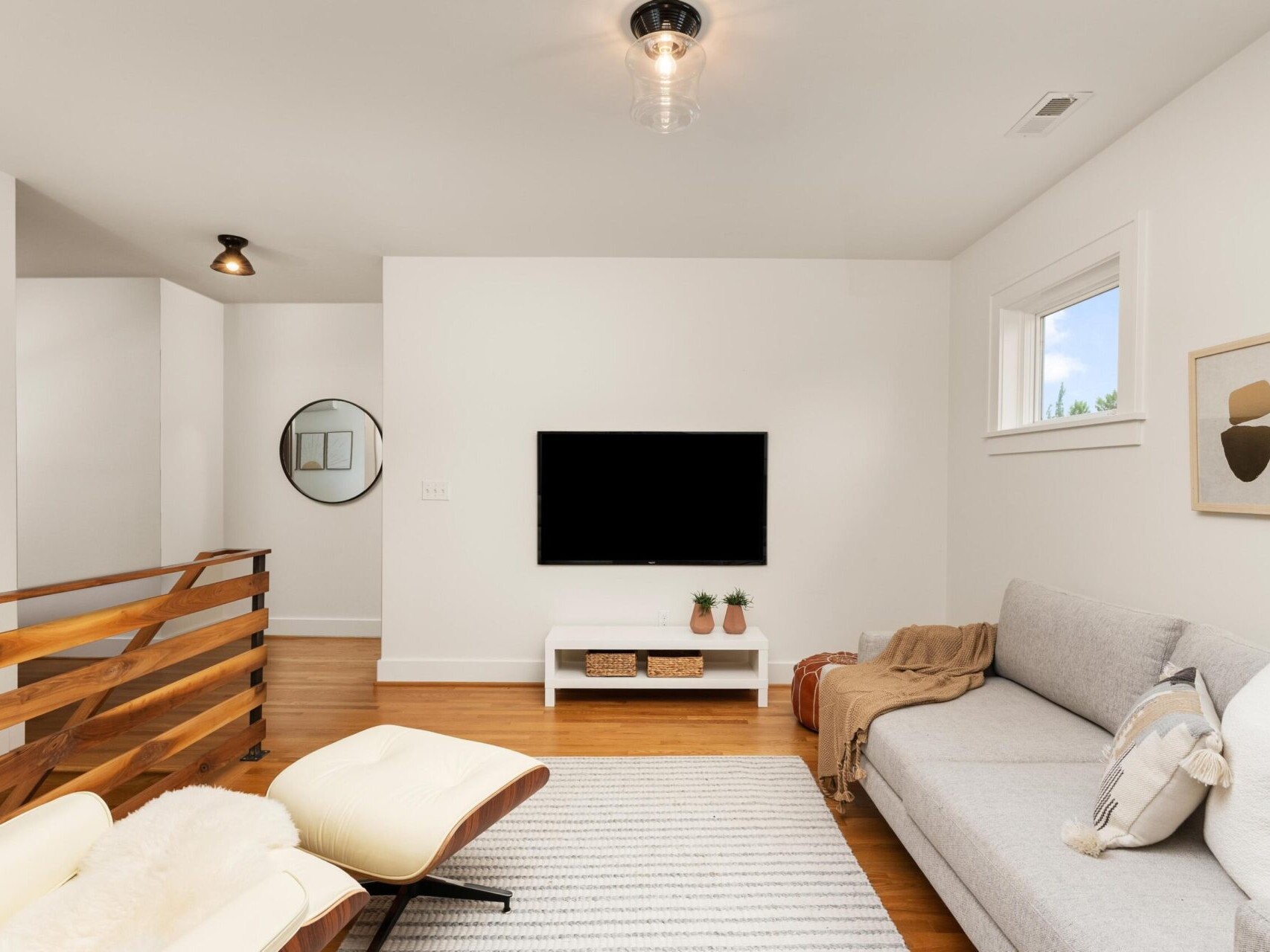Modern living room with white walls featuring a gray sofa, light wood flooring, and a wall-mounted TV. A white and wood lounge chair sits nearby, next to a brown and white railing. A round mirror and minimal decor complete the space.
