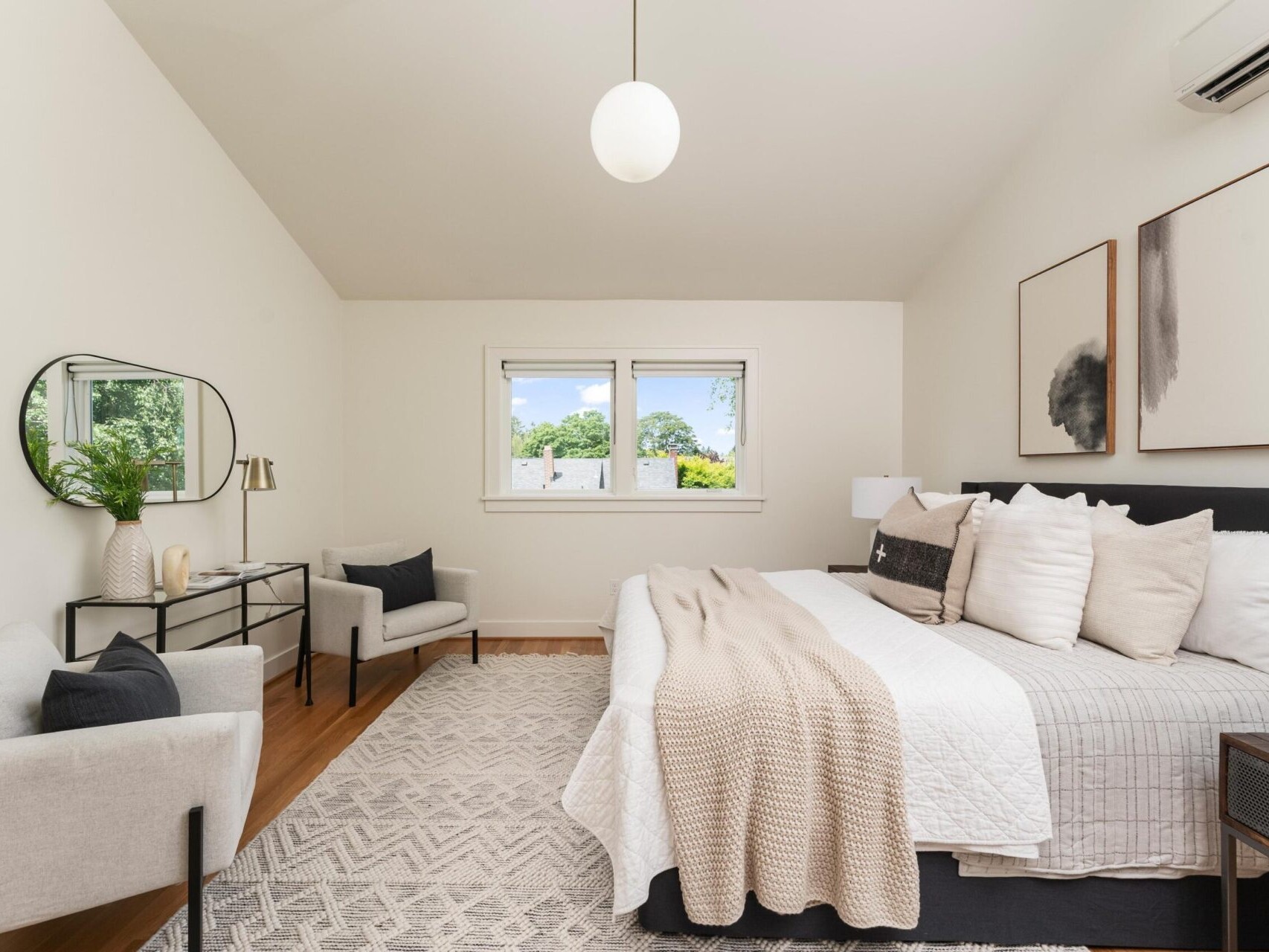 A cozy bedroom with a large bed adorned with white and beige pillows. Two abstract paintings hang above the bed. A round mirror and small decor items sit on a black console. Two chairs flank a small table near a window overlooking greenery.