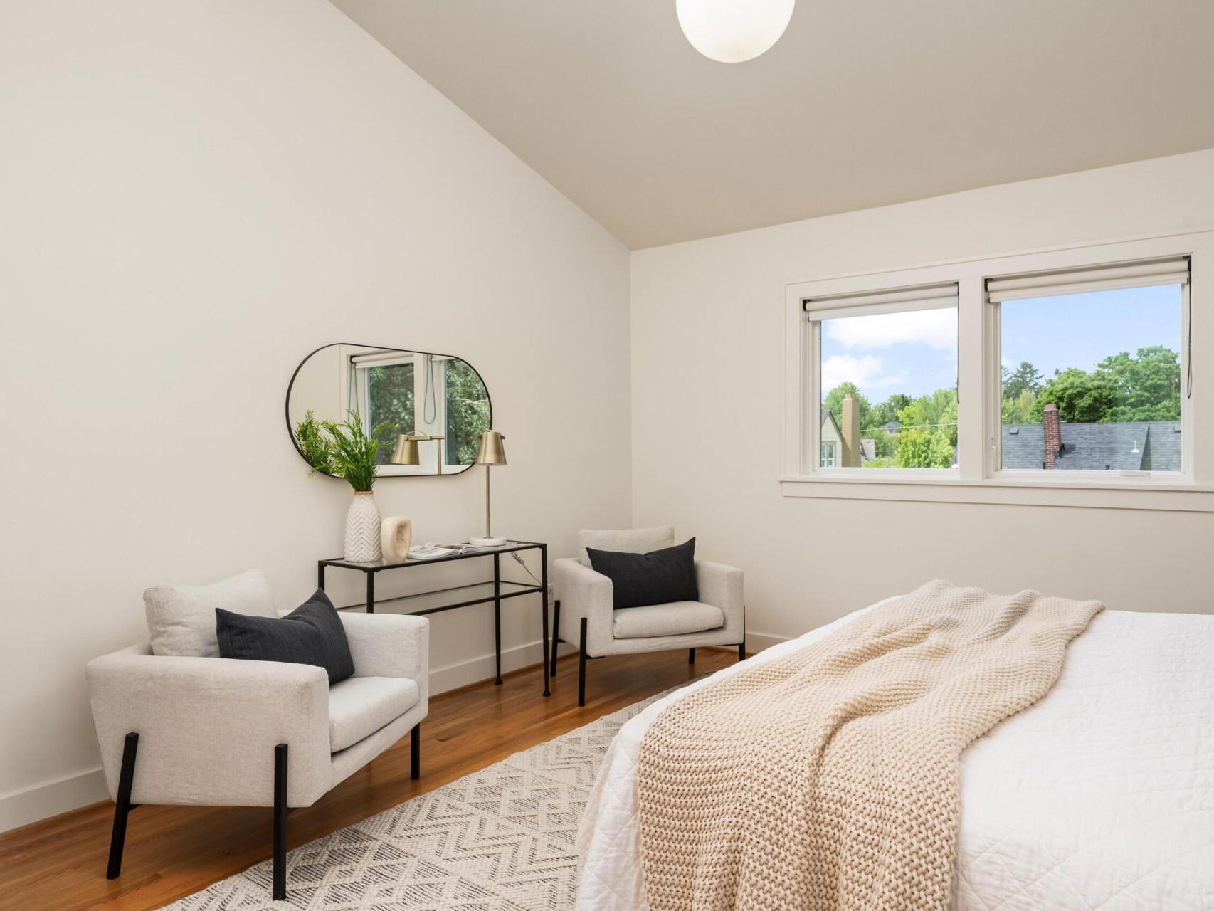 A cozy bedroom with white walls and a sloped ceiling features a bed with a knit throw, two cushioned chairs, and a round mirror above a small table. The window offers a view of trees and rooftops outside.