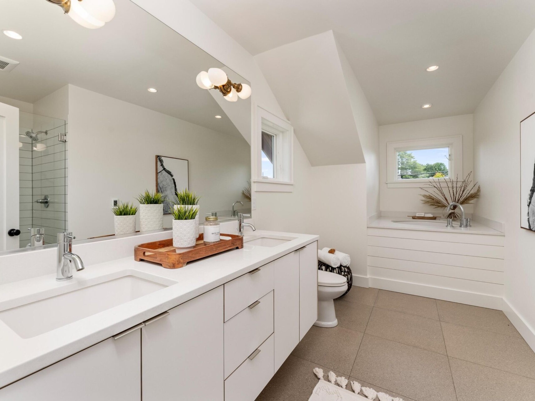 A modern bathroom with a double sink vanity, decorative plants, and a wooden tray. Theres a white toilet, a glass-enclosed shower, and a window above a small ledge with a vase and dried flowers. The room features neutral tones and minimalist decor.