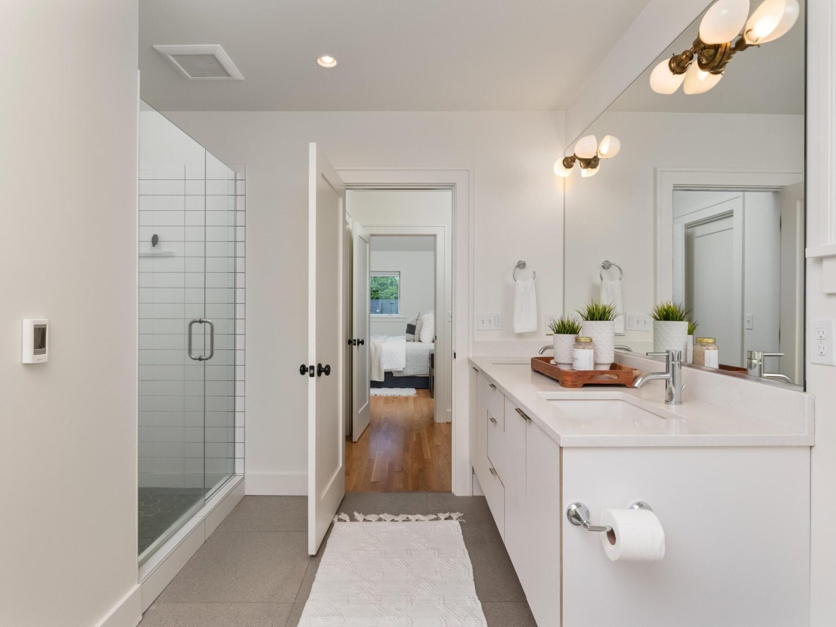 A modern bathroom with a glass-enclosed shower, large mirror, and double sink vanity. The countertop holds a tray with decorative items and plants. A white rug lies on the floor, and the door opens to a bedroom with wooden floors.