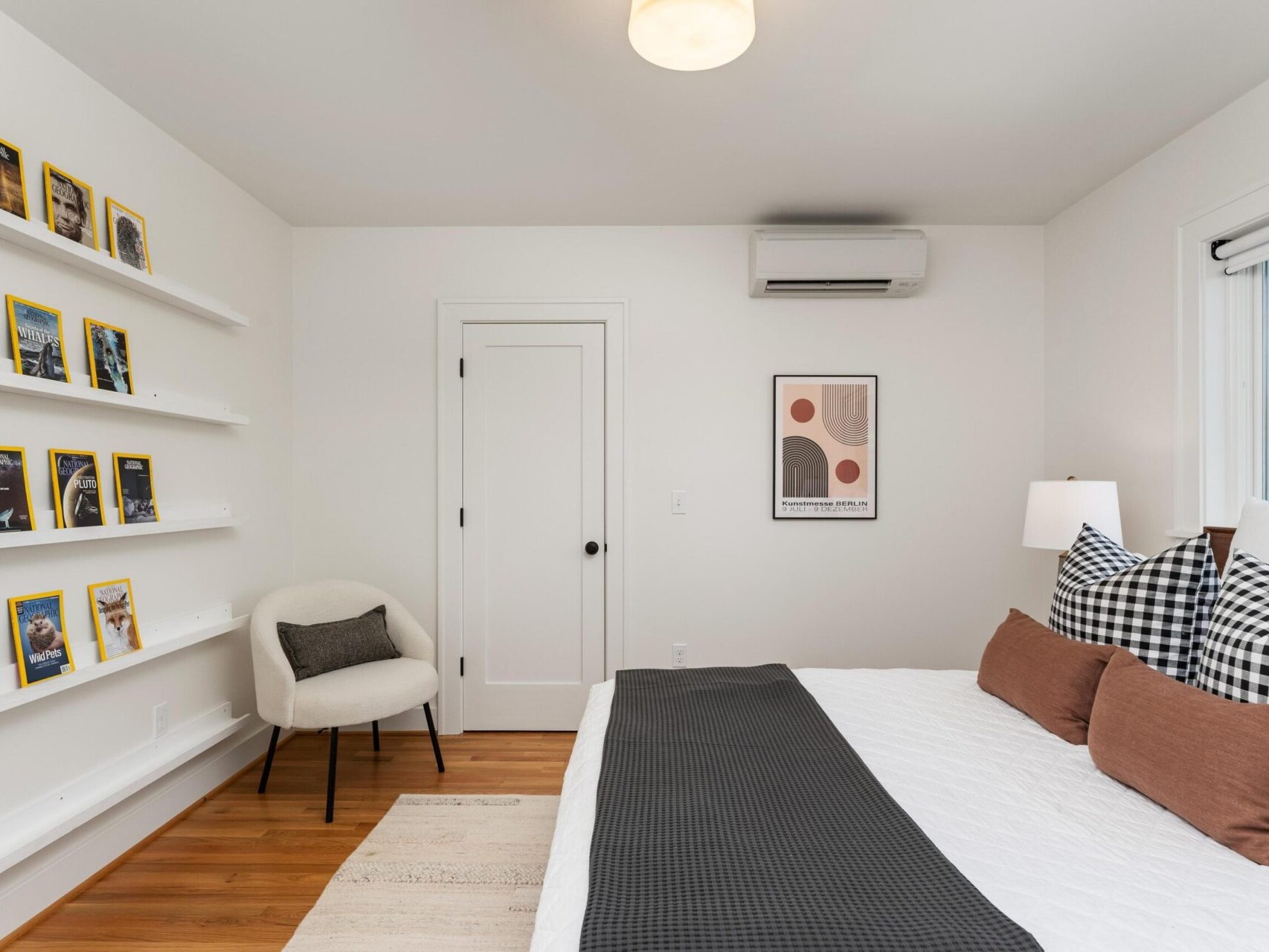 A modern bedroom with a neatly made bed featuring black and white checkered pillows and a dark gray throw. Shelves display yellow-bordered magazines. Theres a white armchair with a brown cushion, a closed door, and a geometric art piece on the wall.