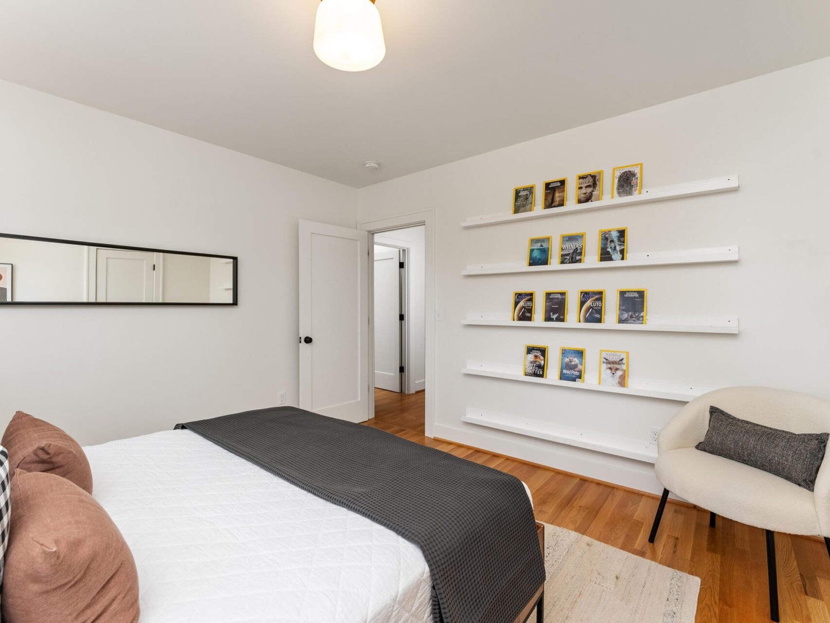 A minimalistic bedroom with a bed featuring black and white pillows and a gray throw. A white armchair sits beside shelves displaying books or frames. A wall mirror is above the bed, and the floor is wooden. The room is well-lit with a ceiling light.