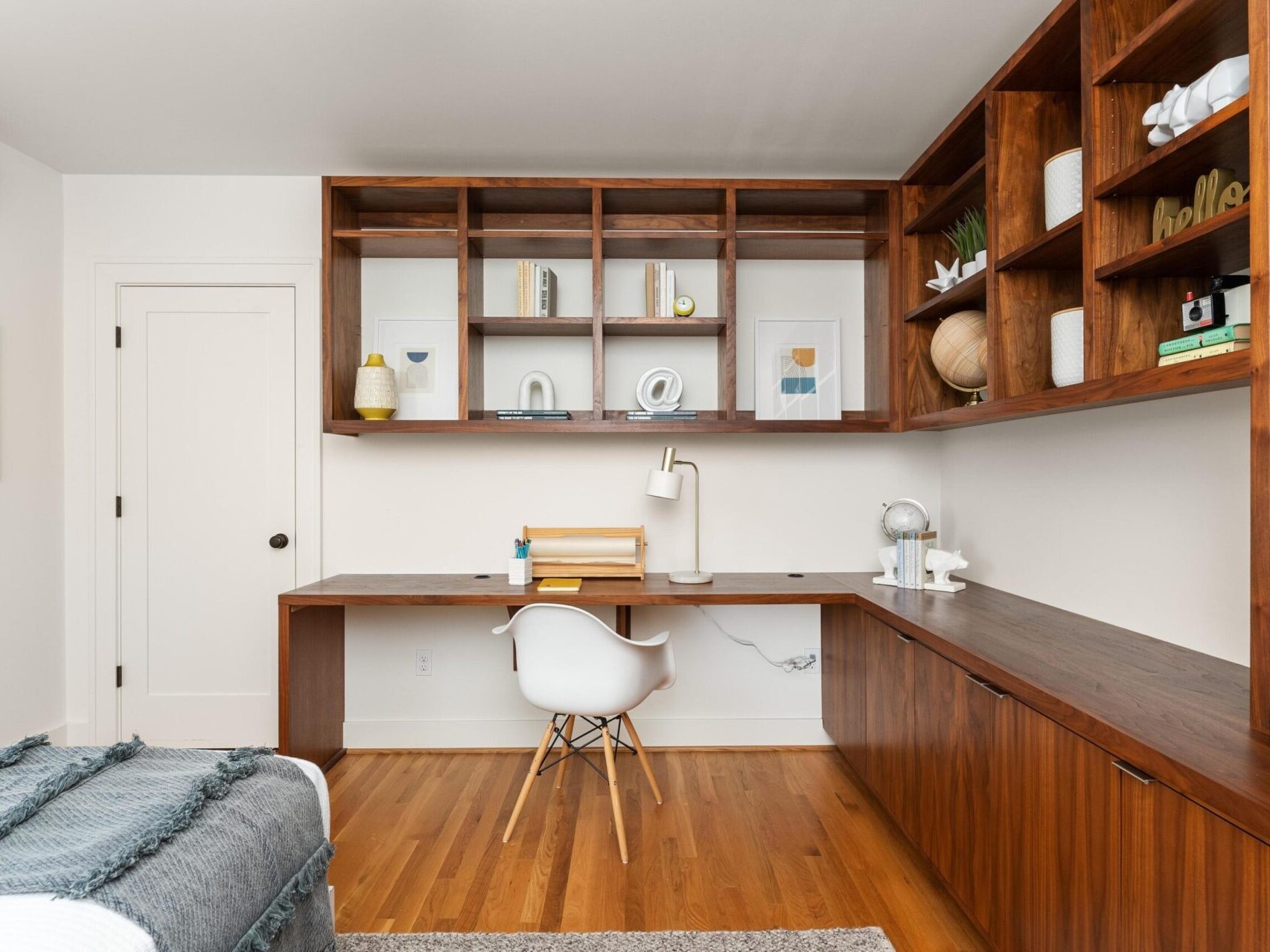 A modern home office with a large wooden built-in desk and shelves. The room features a white chair, decorative objects, books, and plants on the shelves. The floor is hardwood, and theres a cozy blanket draped on a nearby bed.