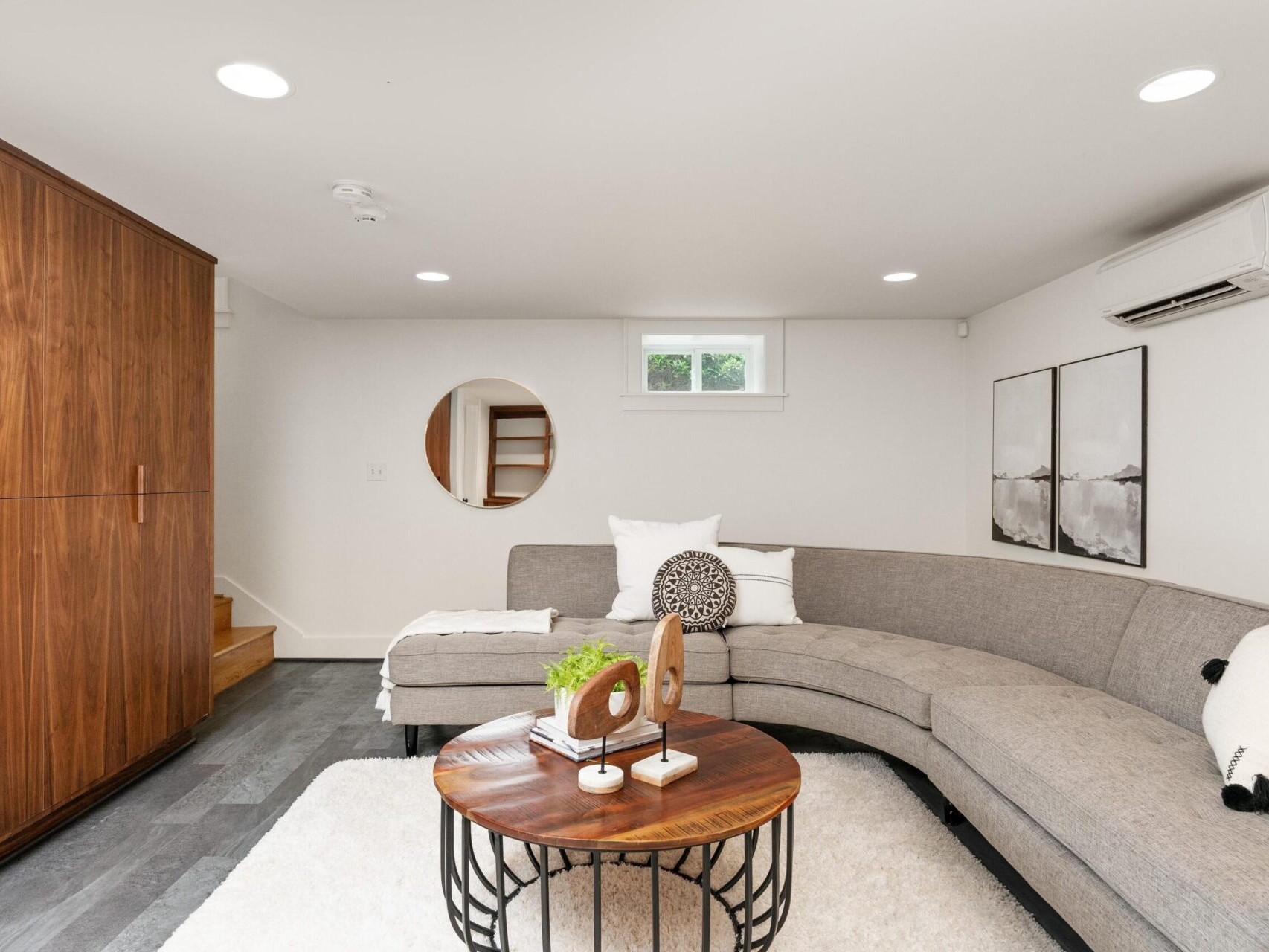 A modern living room features a curved gray sofa with patterned cushions, a round wooden coffee table on a white rug, and abstract wall art. A wooden cabinet is on the left, and a small window and wall-mounted air conditioner complete the space.