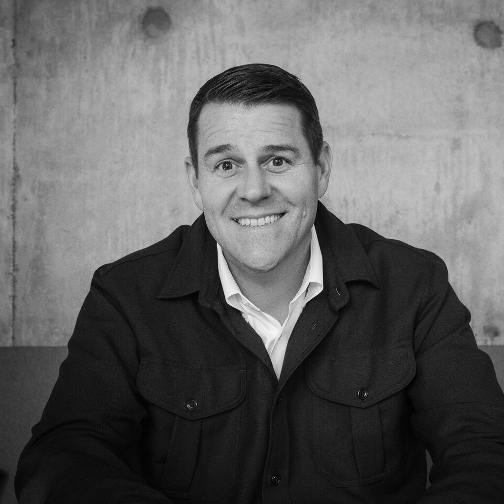 A man wearing a dark jacket and white shirt sits confidently on a couch, smiling at the camera. The background is a textured concrete wall, lending a classic feel to this black and white image of a top real estate agent from Portland, Oregon.