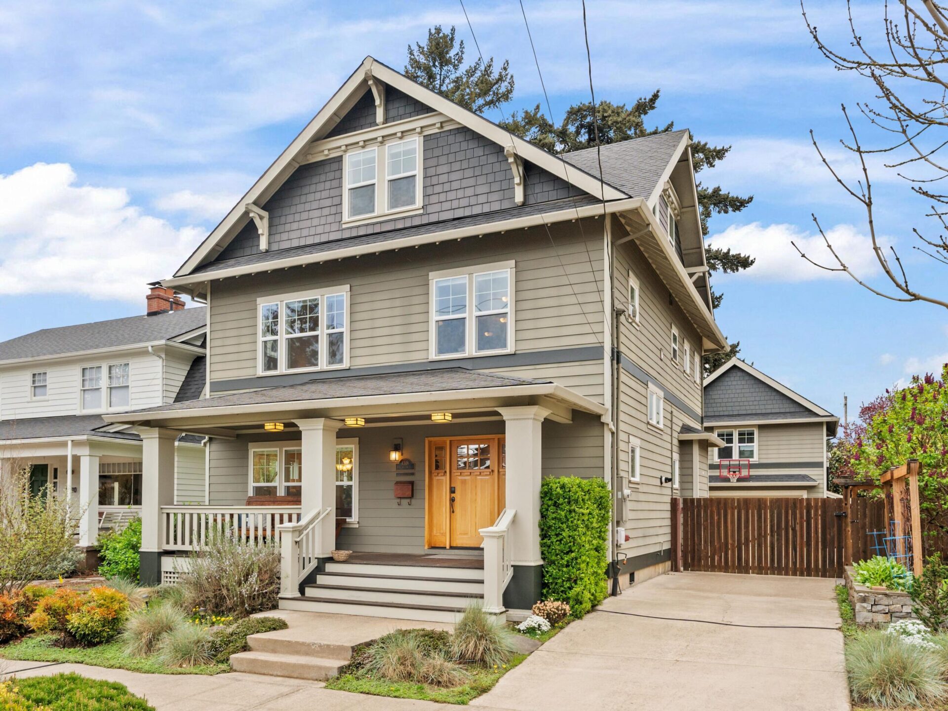 A two-story traditional house in Portland, Oregon real estate with gray siding, a gabled roof, and a welcoming porch. A wooden front door is flanked by windows. There's a driveway on the right and a lush garden with shrubs in the front. Tall trees are in the background.