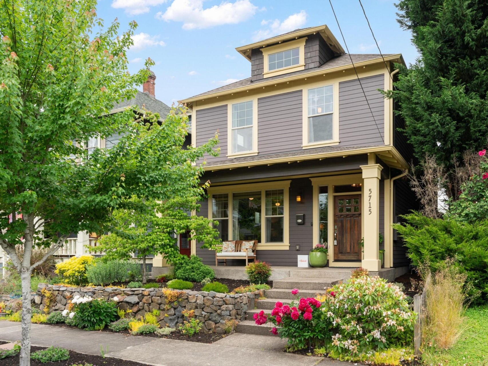 A charming two-story house with brown siding and cream trim, featuring a porch with seating. Nestled in Portland's vibrant market, this gem is surrounded by a well-maintained garden and stone retaining wall, set against tall trees and a clear sky—a dream home for any discerning Portland Oregon real estate seeker.