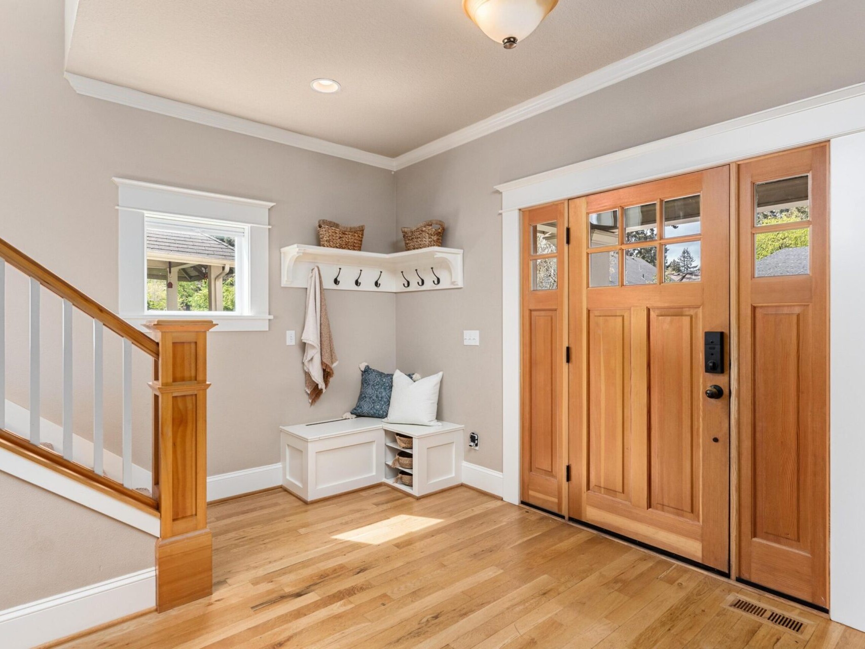 A bright and airy entryway featuring light wood flooring, a wooden staircase with white accents, and a wooden front door. A built-in bench with storage and hooks sits against a light-colored wall under a small window. Discover your dream home in Portland Oregon Real Estate today.