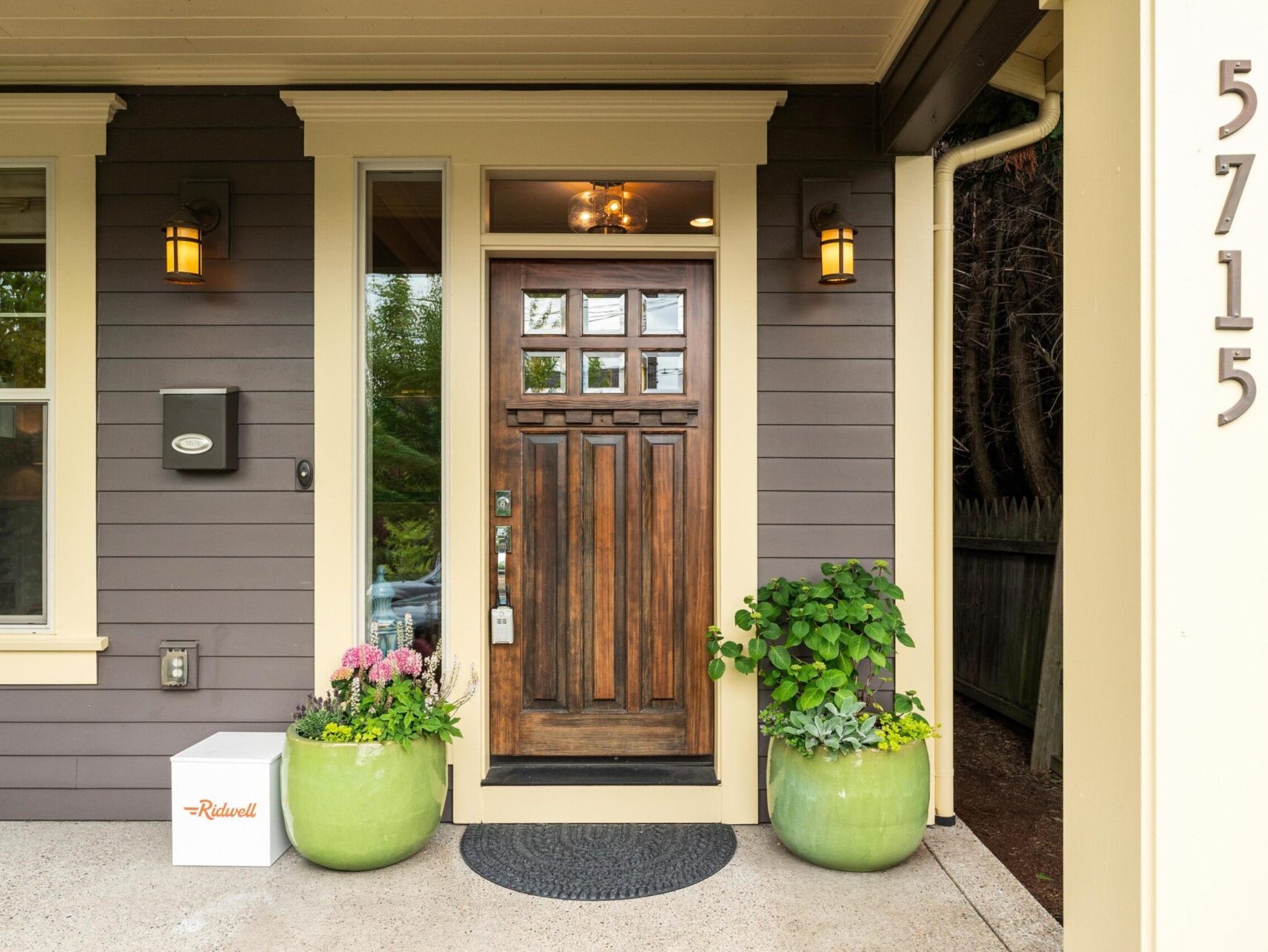 Cozy front entrance of a Portland home with a wooden door, flanked by green potted plants and a white delivery box labeled Redwood. The house number 5715 decorates a beige pillar, while outdoor lights add charm. Find your dream house today with the top Portland Realtor.