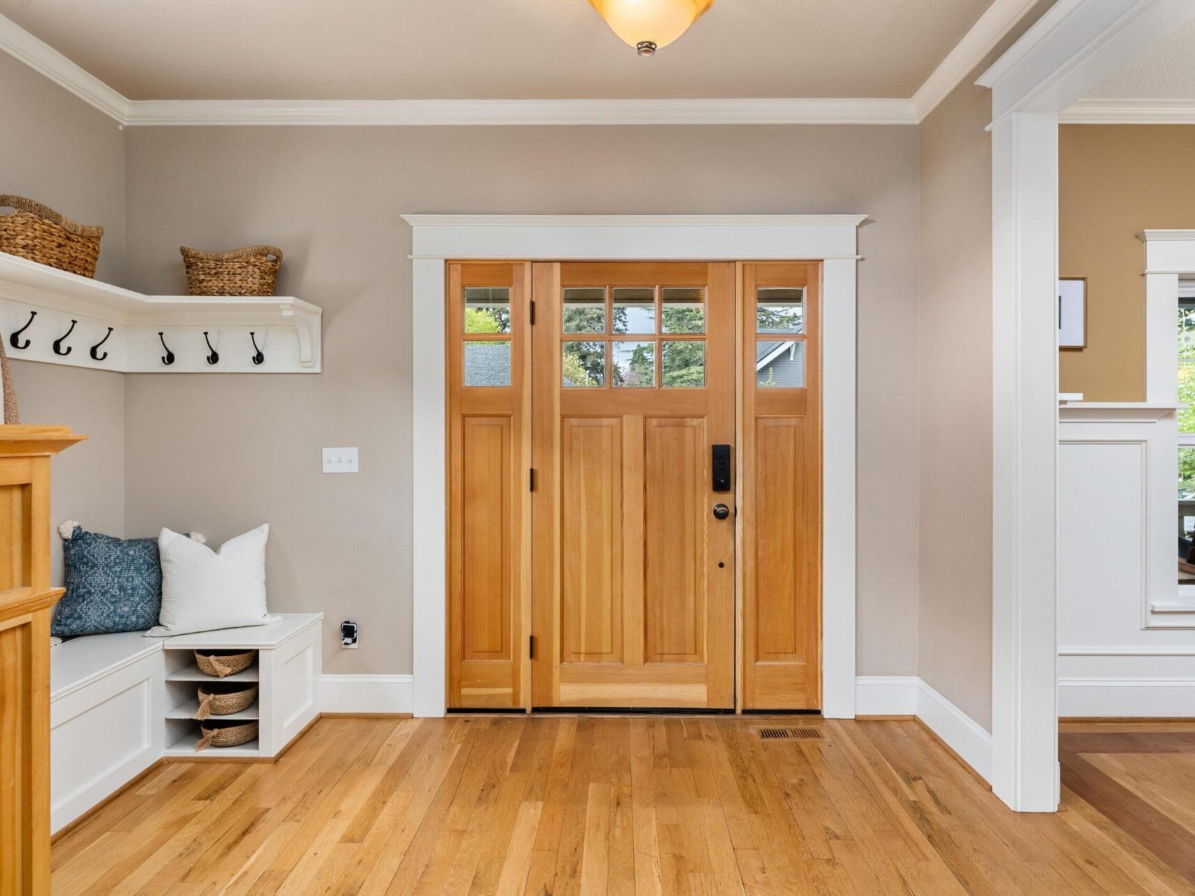 A welcoming entryway with wooden double doors, a bench adorned with blue and white pillows, coat hooks, and wicker baskets on a shelf awaits you. Natural light illuminates the hardwood floor, creating a warm and inviting atmosphere—a quintessential Portland Oregon real estate gem.