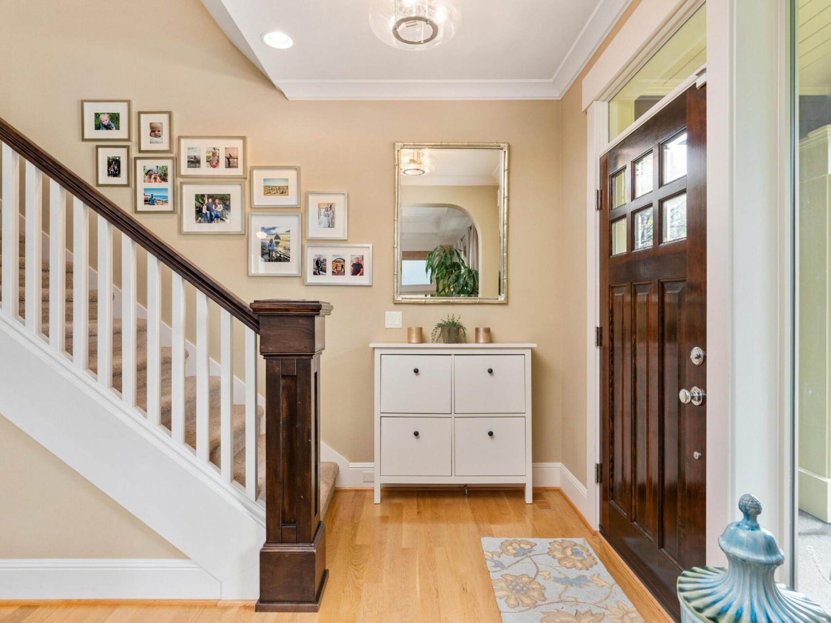 A welcoming foyer in Portland Oregon Real Estate, featuring a wooden front door, light wood flooring, and a staircase with white railings. The wall is adorned with family photos and a mirror above a white storage cabinet. A decorative blue vase sits on the floor.