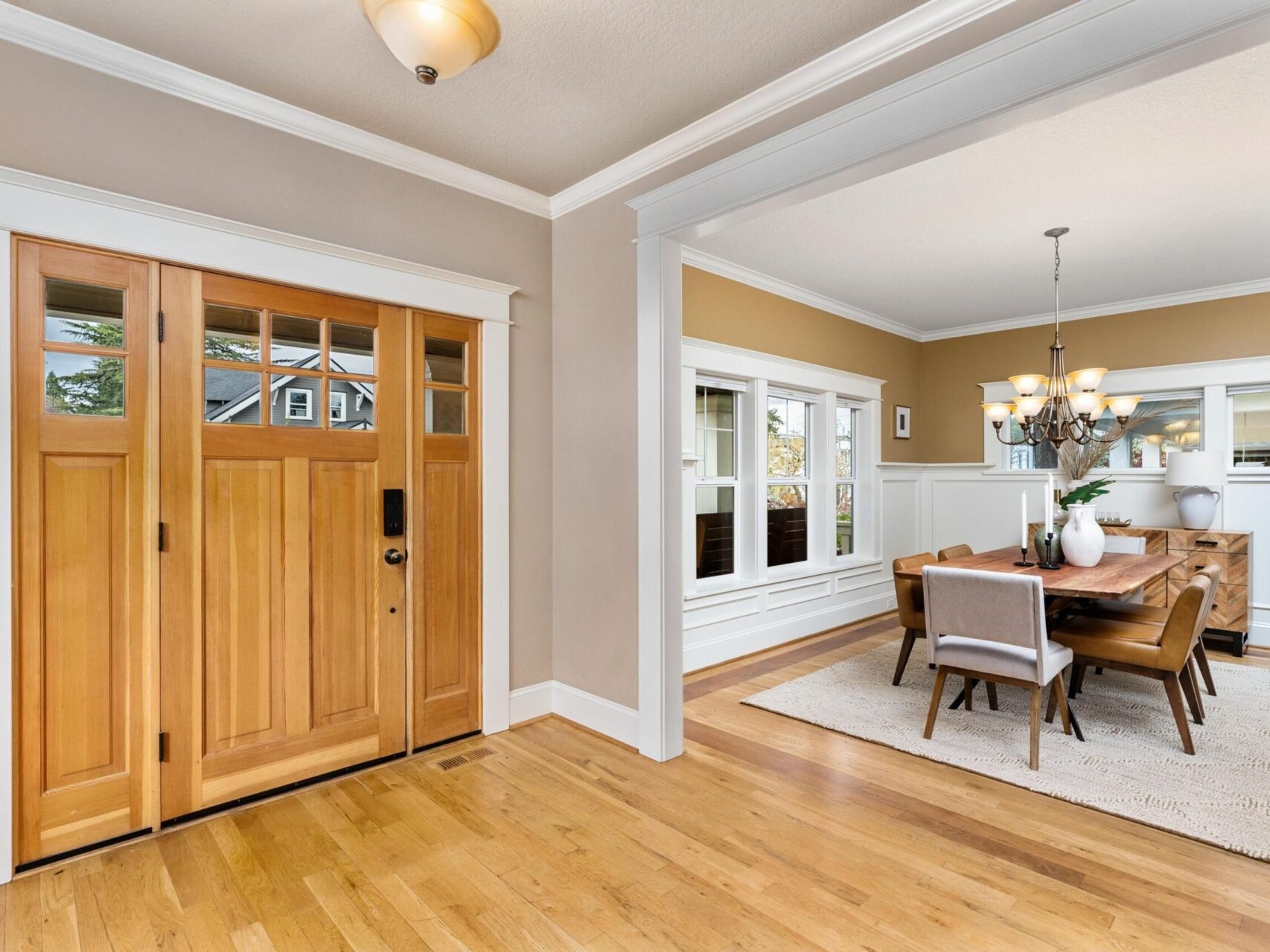 A warm, inviting entryway with wooden double doors opens into a dining room. The dining area boasts a wooden table, cushioned chairs, and a chandelier. Neutral walls and ample windows provide a bright, airy atmosphere⁠—an ideal space highlighted by top Portland Realtor expertise.