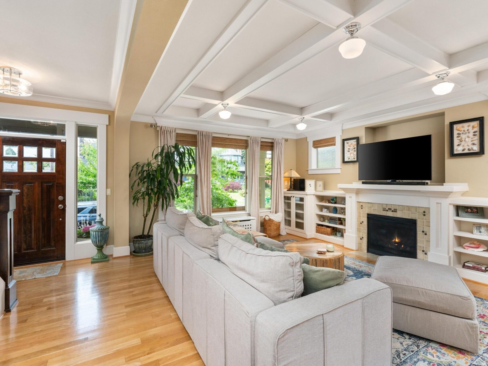 A cozy living room with a large gray sectional sofa facing a TV above a fireplace, perfect for those exploring Portland Oregon real estate. It features wooden floors, a patterned rug, and large windows with beige curtains. Potted plants add greenery, and there's a staircase leading upstairs.