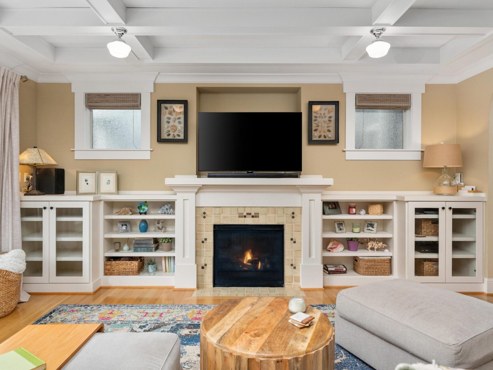 A cozy living room in a Portland home showcases beige walls, featuring a fireplace beneath a wall-mounted TV. Built-in shelves flank the fireplace with books and decor. A colorful rug, round wooden coffee table, and light gray seating complete the space—a gem for any Portland Realtor to highlight.