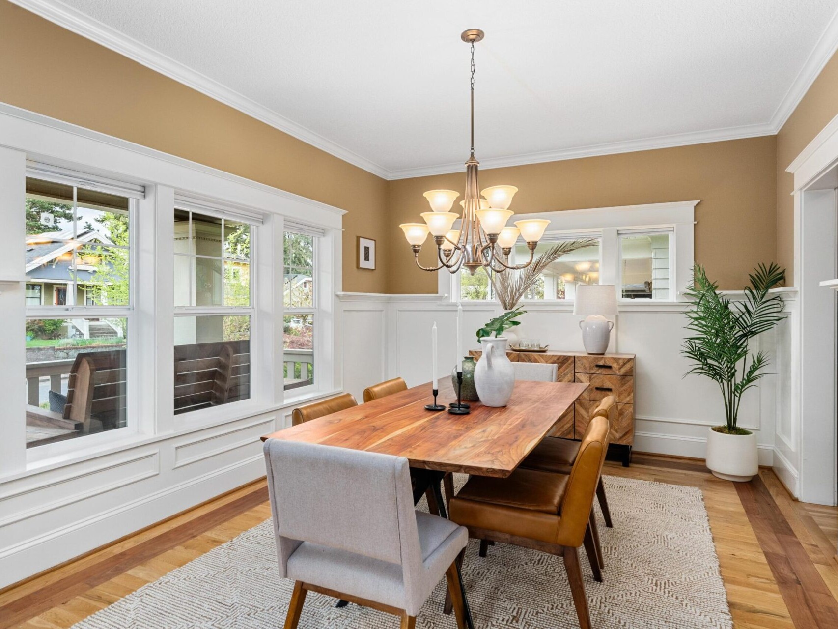 A dining room with a wooden table and six chairs sits gracefully under a chandelier, surrounded by elegant white wainscoting and beige walls. Large windows flood the space with light, while the sideboard and potted plant add charm—perfect for those exploring Portland Oregon real estate.