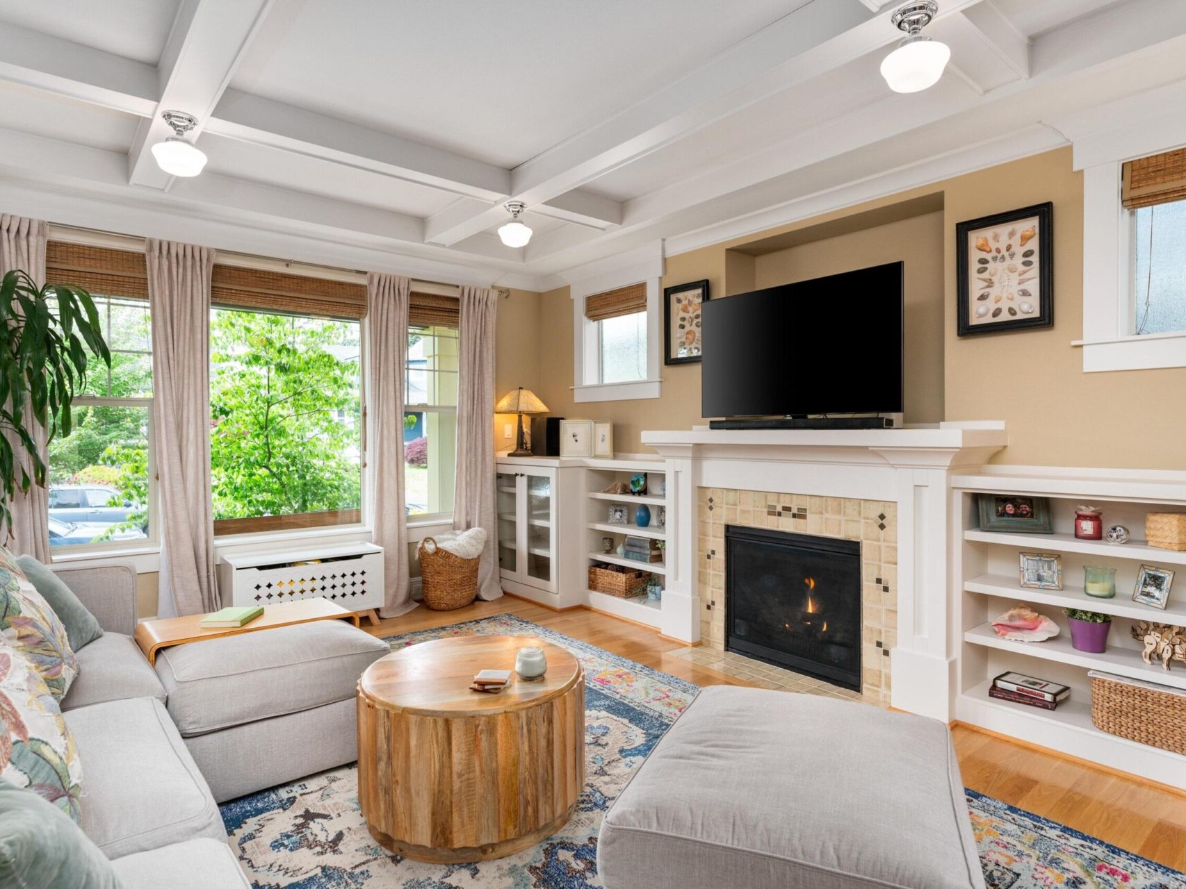 A cozy living room in Portland Oregon real estate, featuring a gray sectional sofa, a round wooden coffee table, and a patterned rug. A fireplace with a TV above is flanked by built-in shelves. Large windows bring in natural light, and potted plants add greenery.