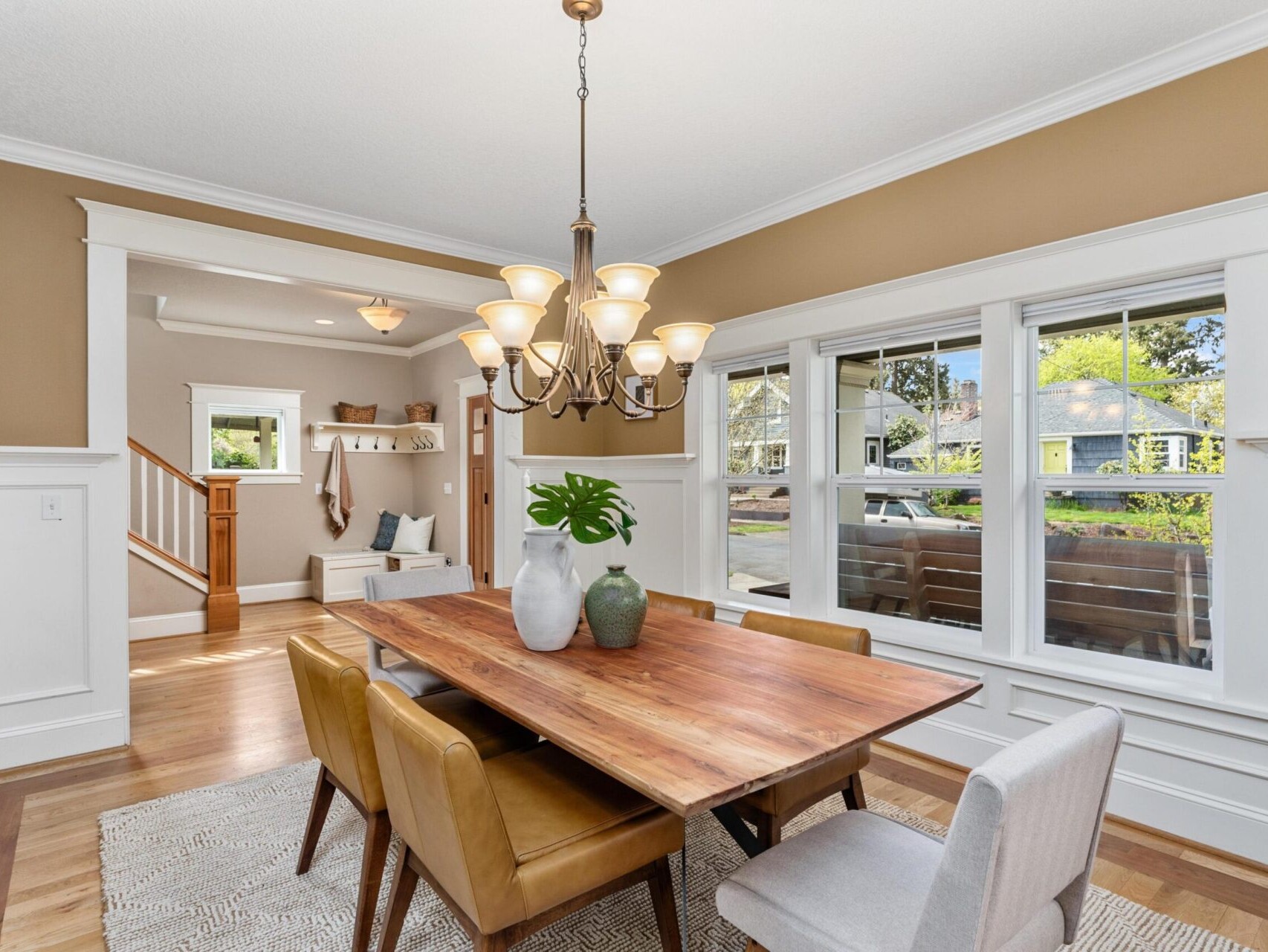 A dining room in Portland Oregon real estate, with a wooden table and six chairs, featuring a chandelier above. A cozy nook with a bench is visible by stairs. Large windows let in natural light, highlighting the neutral tones and greenery on the table.