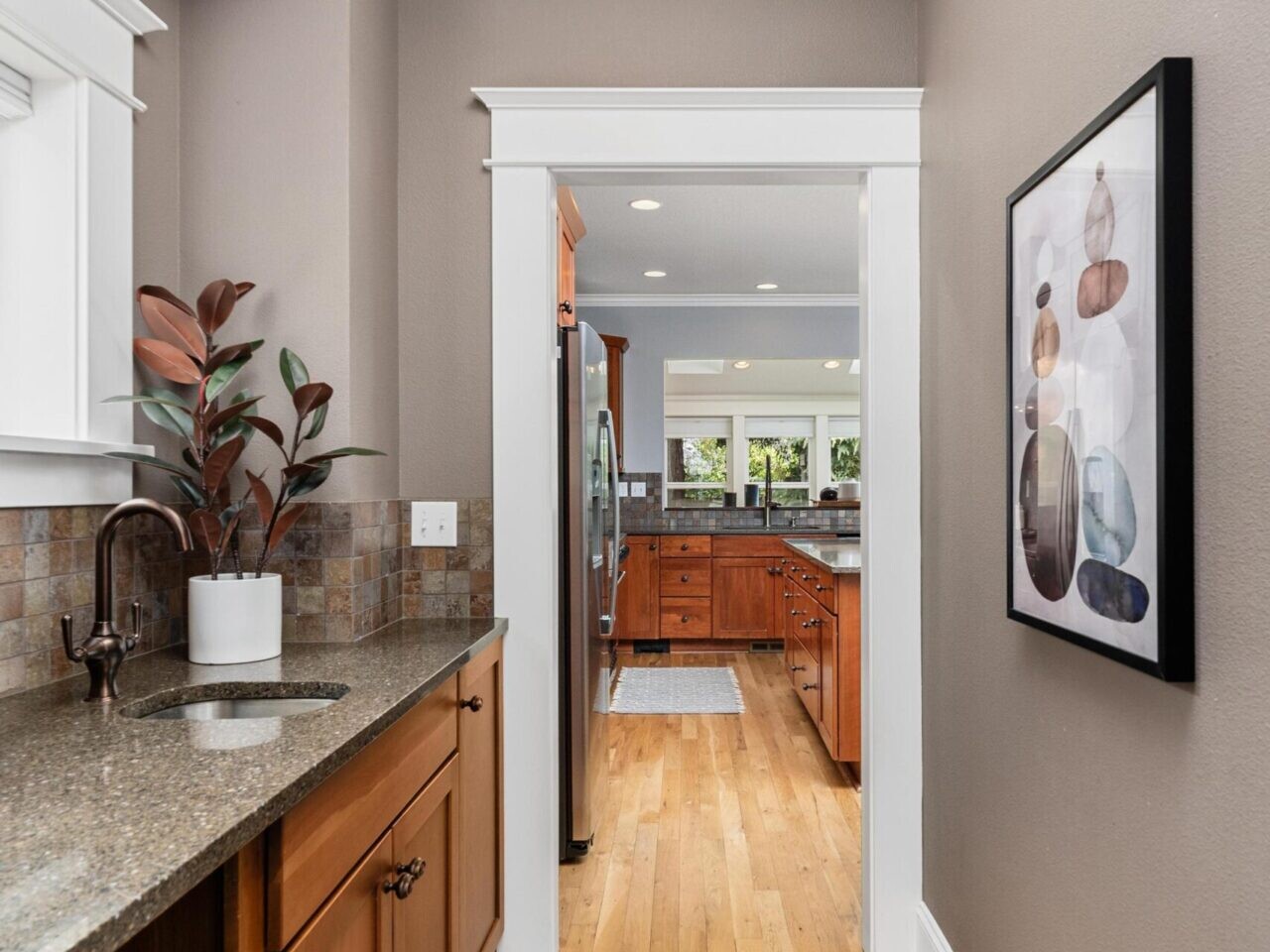 Narrow kitchen with wood cabinets, granite countertops, and a tile backsplash on the left. Wooden floors lead to a wider area with more cabinets and stainless steel appliances. Framed artwork hangs on the right wall. Brightly lit and modern, this gem is a highlight in Portland Oregon real estate.