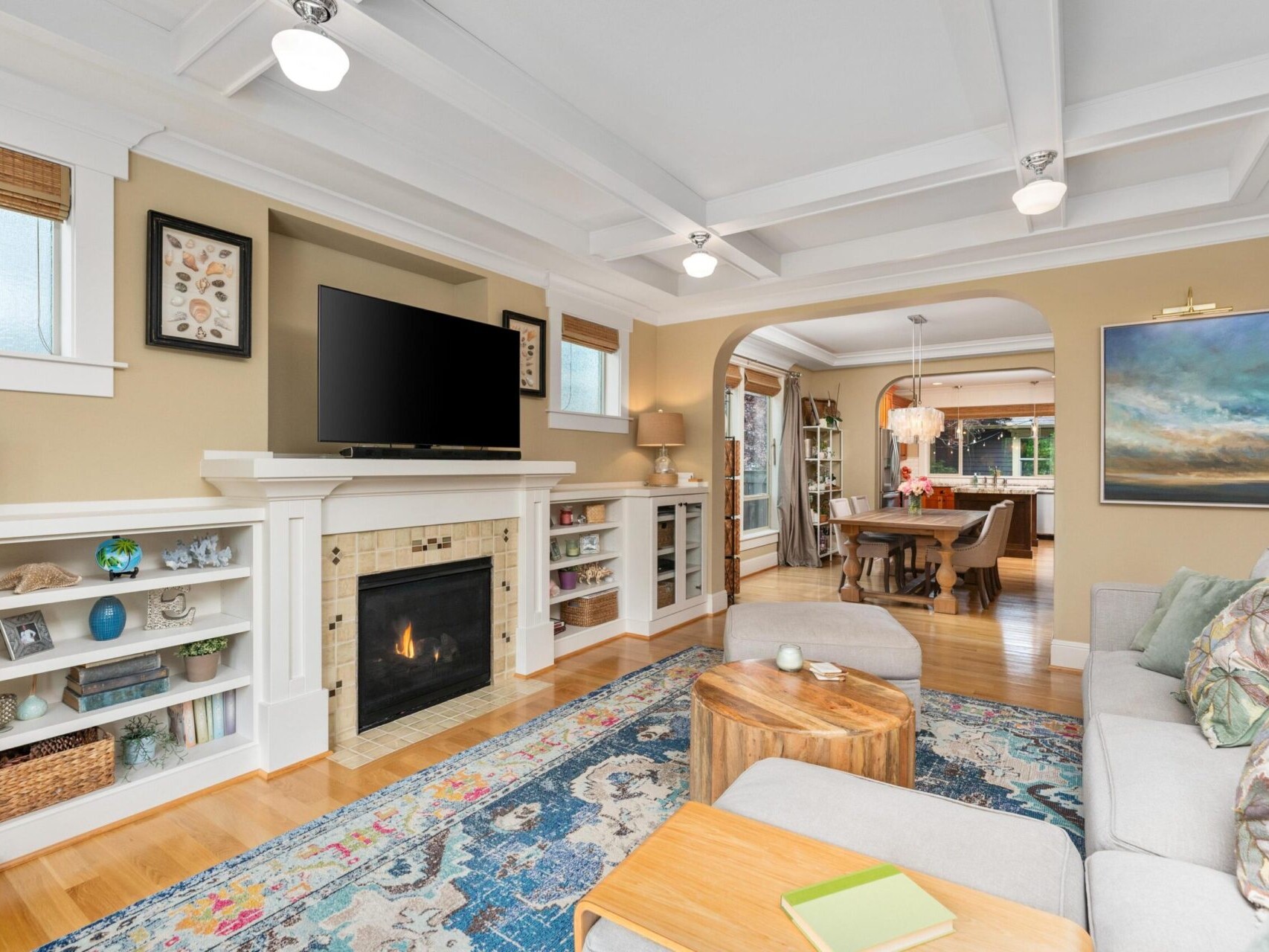 A cozy living room in Portland, Oregon, features a light gray sofa, colorful rug, and framed art on beige walls. The fireplace with built-in shelves holds decor items. A large TV sits above the fireplace, while an archway leads to the dining area with a wooden table—a gem in Portland real estate.