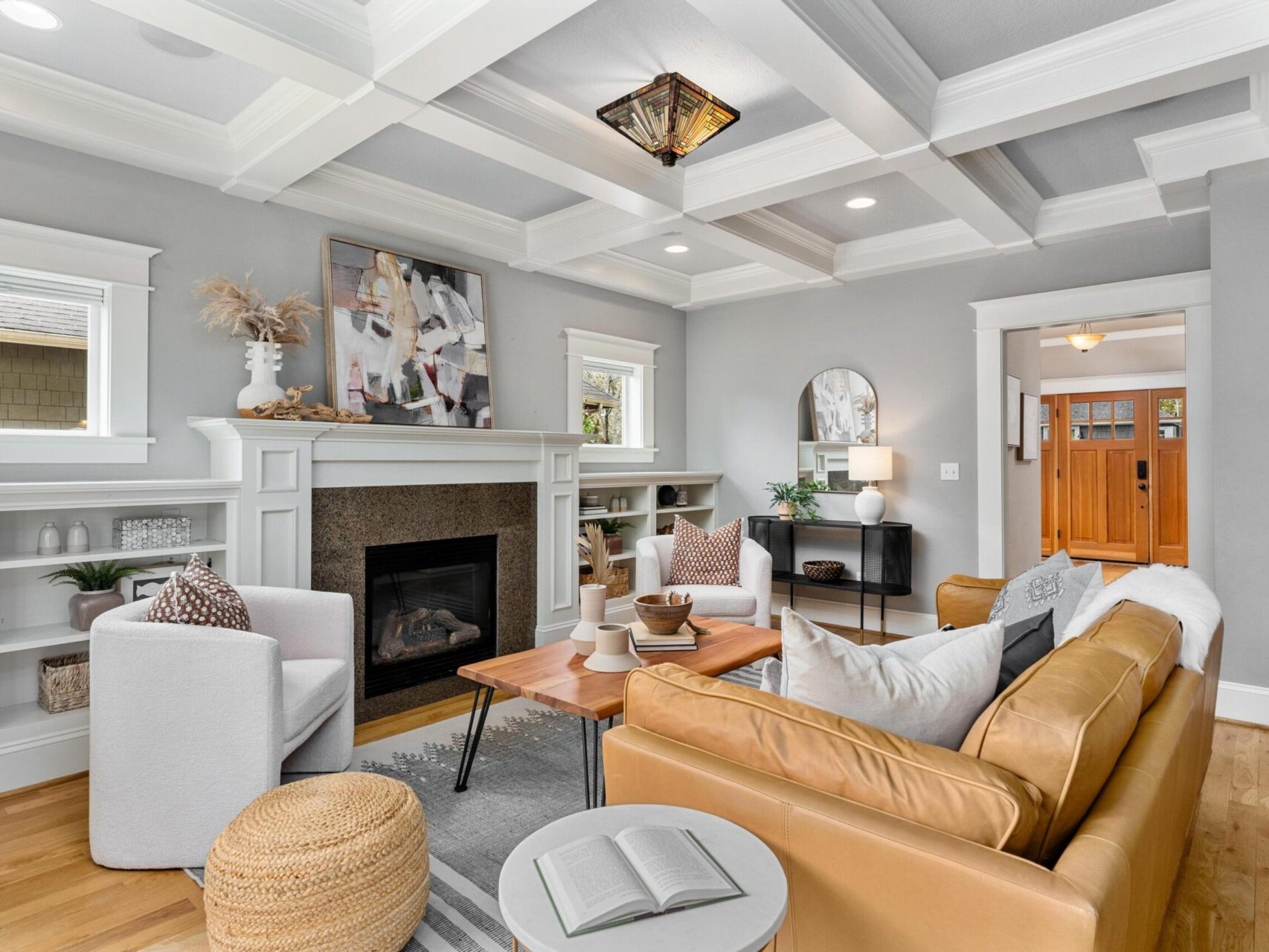 A bright, stylish living room in a Portland, Oregon real estate gem features a tan leather sofa, a wooden coffee table, and a modern armchair. The space includes a cozy fireplace, built-in shelves, and artistic decor. Natural light streams through the windows, enhancing the airy feel.
