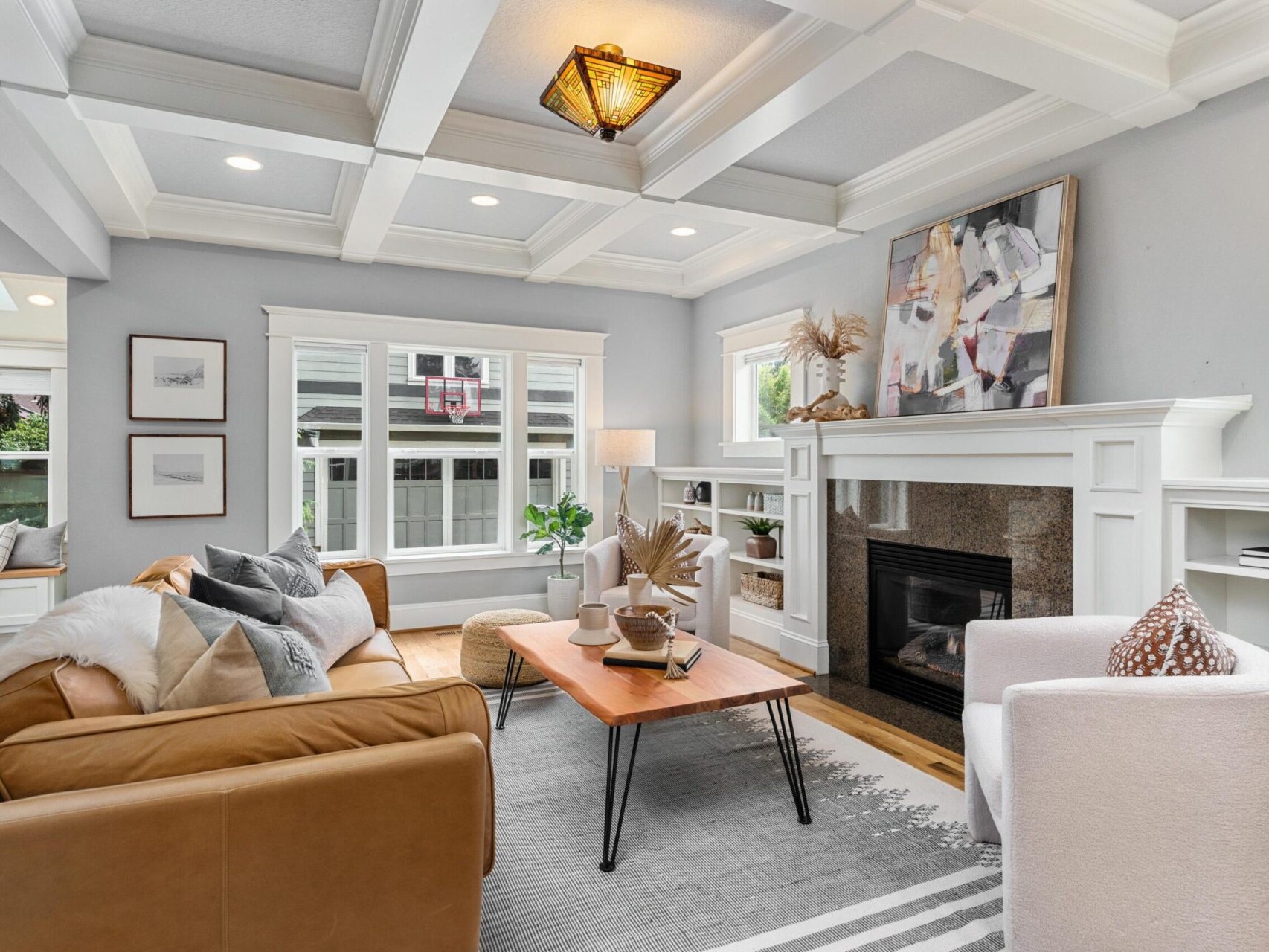 A cozy living room in a Portland Oregon real estate listing features a brown leather sofa, white armchair, and wooden coffee table on a gray rug. A fireplace with a painting above it is on the right, and windows allow natural light to fill the space. Built-in shelves decorate the walls.