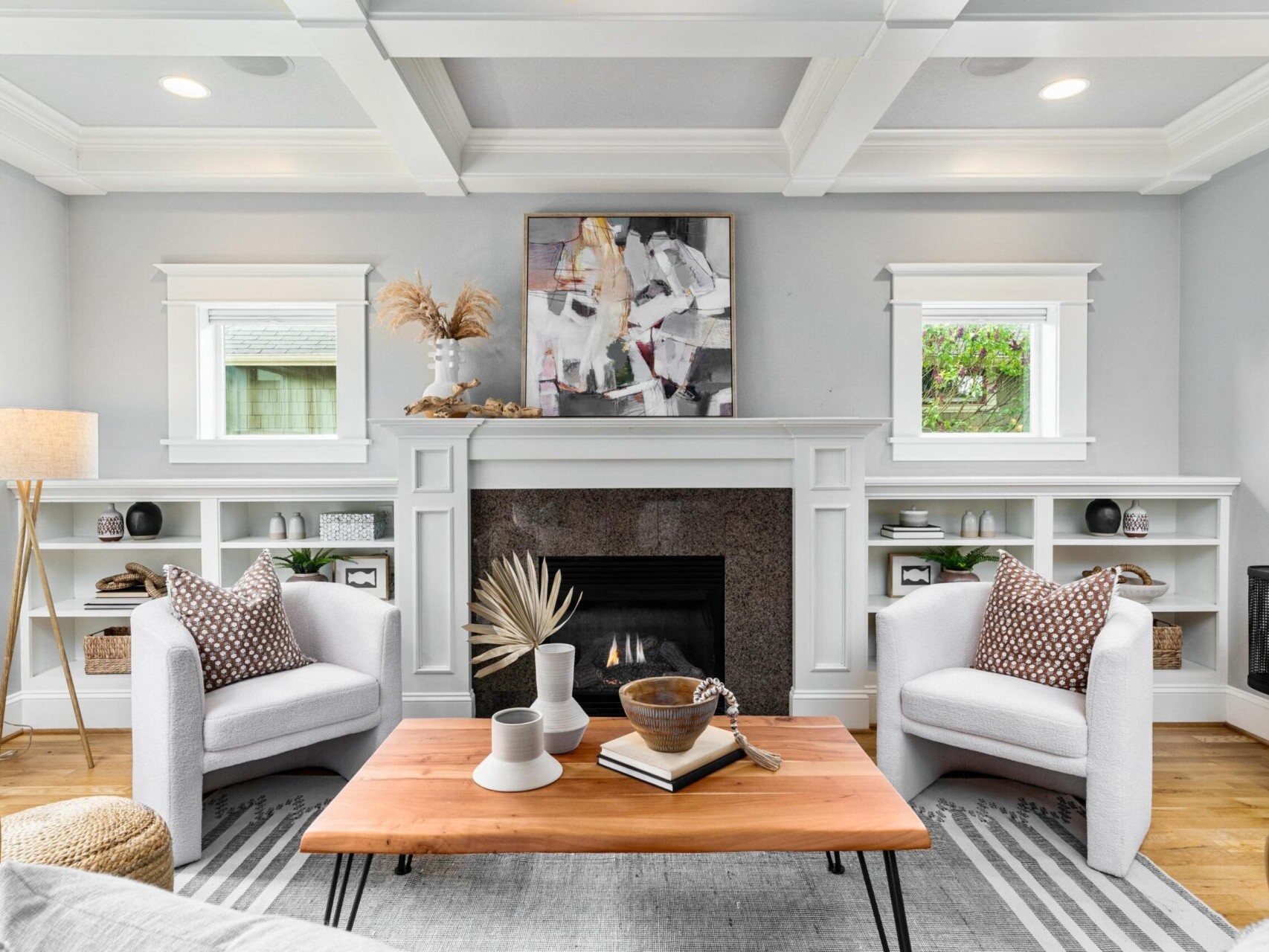 A cozy living room in Portland Oregon Real Estate features two white armchairs facing a wooden coffee table. A fireplace with a dark stone surround is nestled between built-in shelves. A modern abstract painting hangs above, illuminated by soft natural light streaming through the windows.