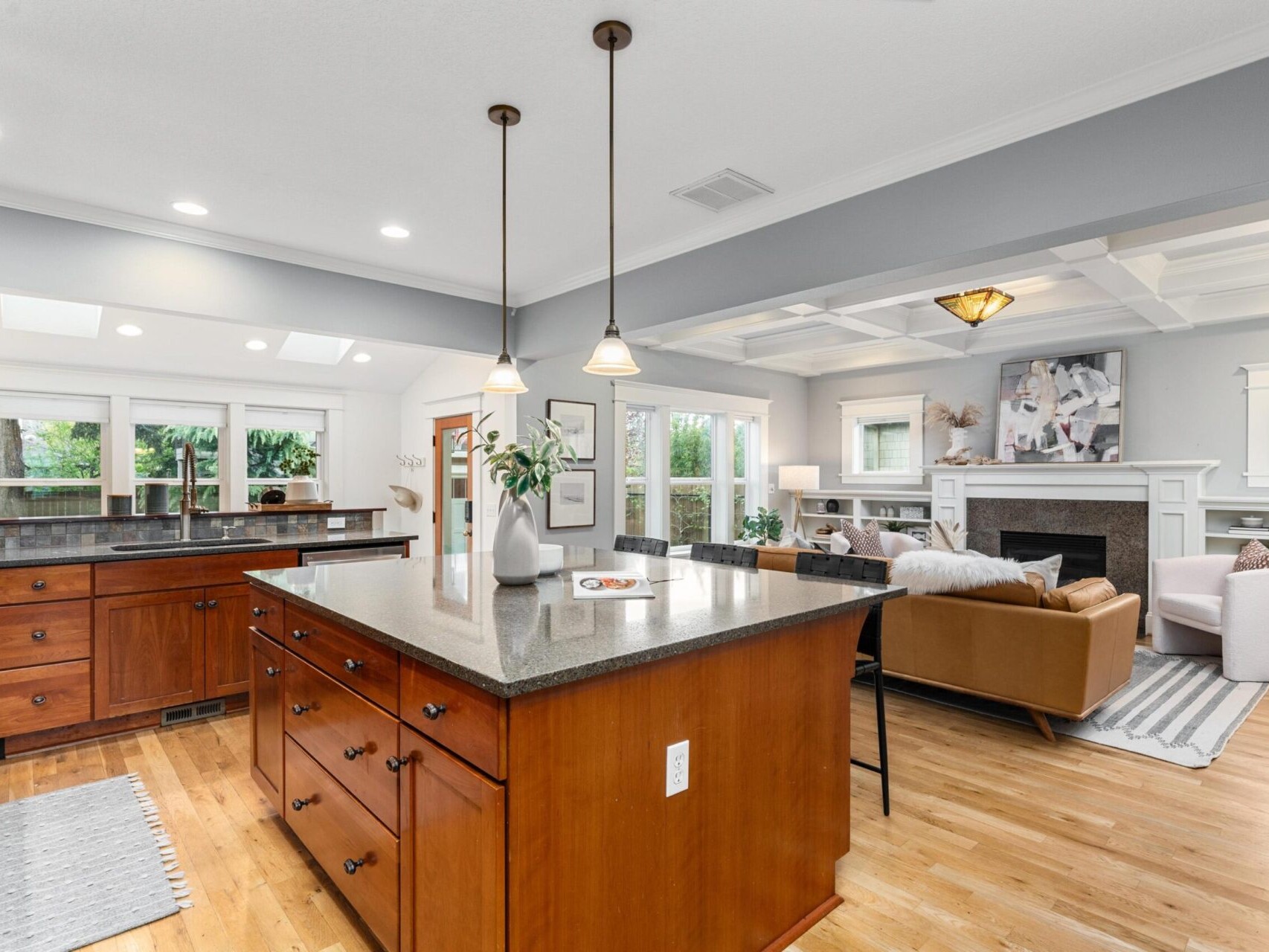 Discover this spacious open-concept kitchen and living room with wooden flooring in the heart of Portland, Oregon real estate. The kitchen features a large central island with pendant lights, while the living room boasts a brown sofa, a white fireplace, and large windows bathing the space in natural light.