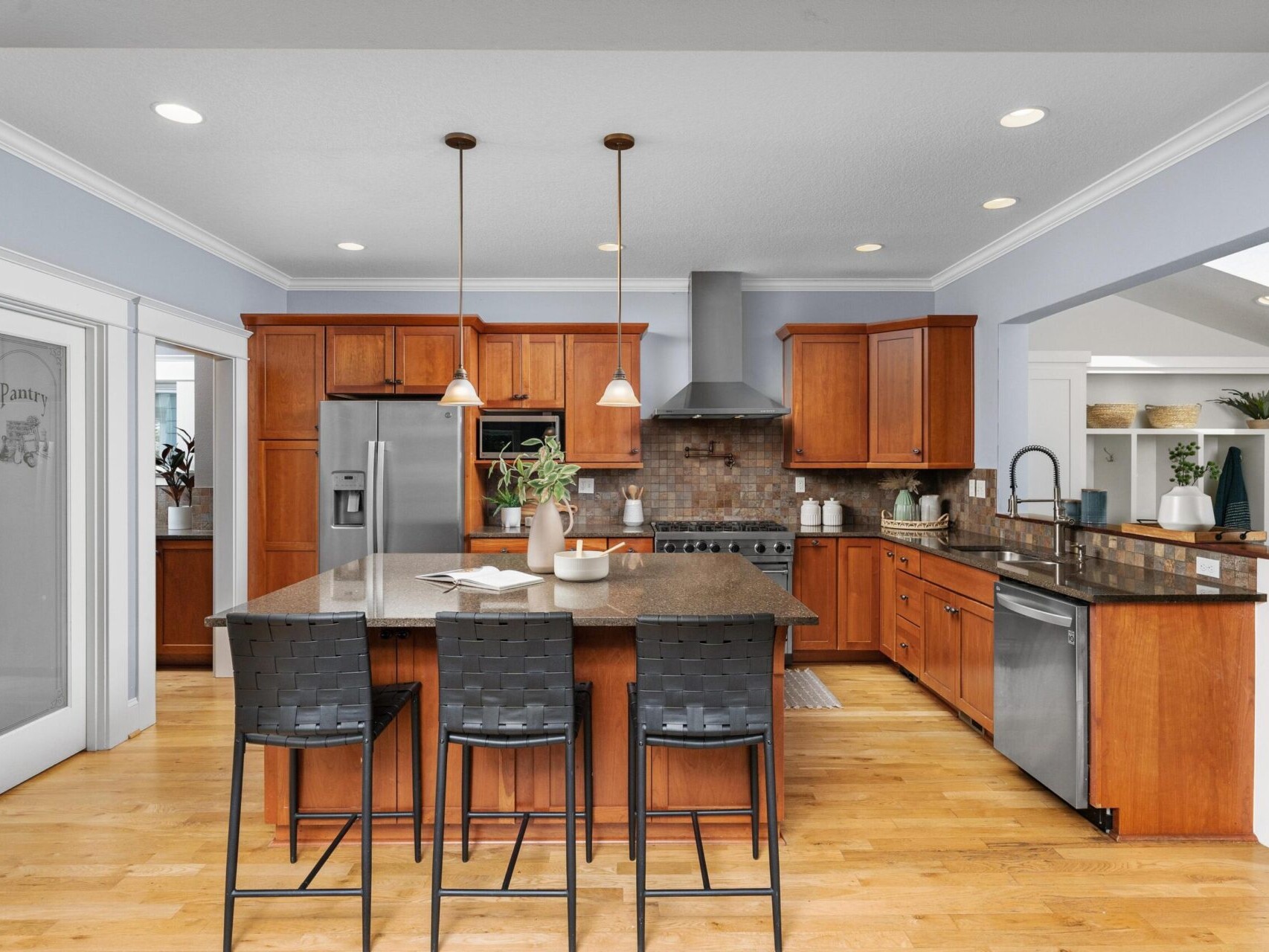 A modern kitchen with wooden cabinets, a granite island, and three black stools. Stainless steel appliances include a fridge and stove. Two pendant lights hang above the island. This bright, open space features light wooden floors and a pantry door—perfect for Portland Oregon real estate seekers.