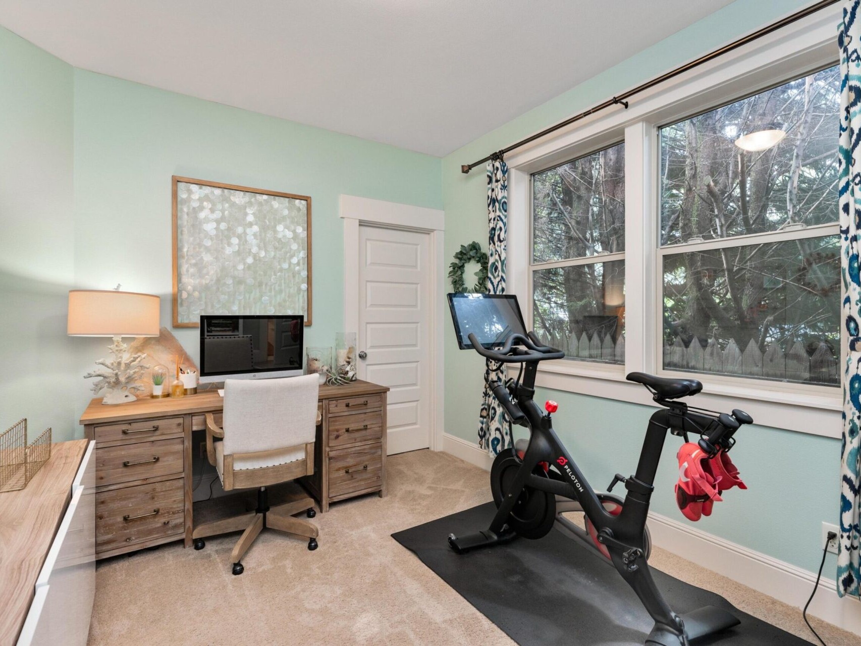 A home office with a wooden desk and computer near a large window covered by blue and white curtains is perfect for productivity. A stationary exercise bike sits nearby on a mat, complementing the room's light green walls and beige carpet—a setup favored by top Portland real estate agents.