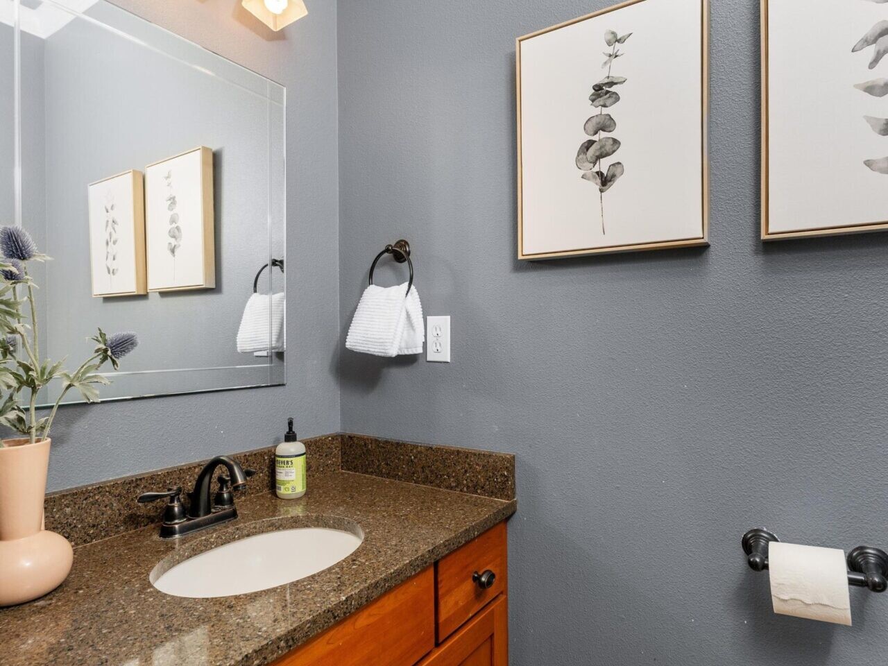 In a cozy bathroom often seen in Portland Oregon real estate, you'll find a brown granite countertop with a wooden vanity, round sink, and black faucet. A mirror sits above the sink, flanked by multi-bulb lighting. Two botanical prints enhance the gray walls, with a towel neatly hung nearby.