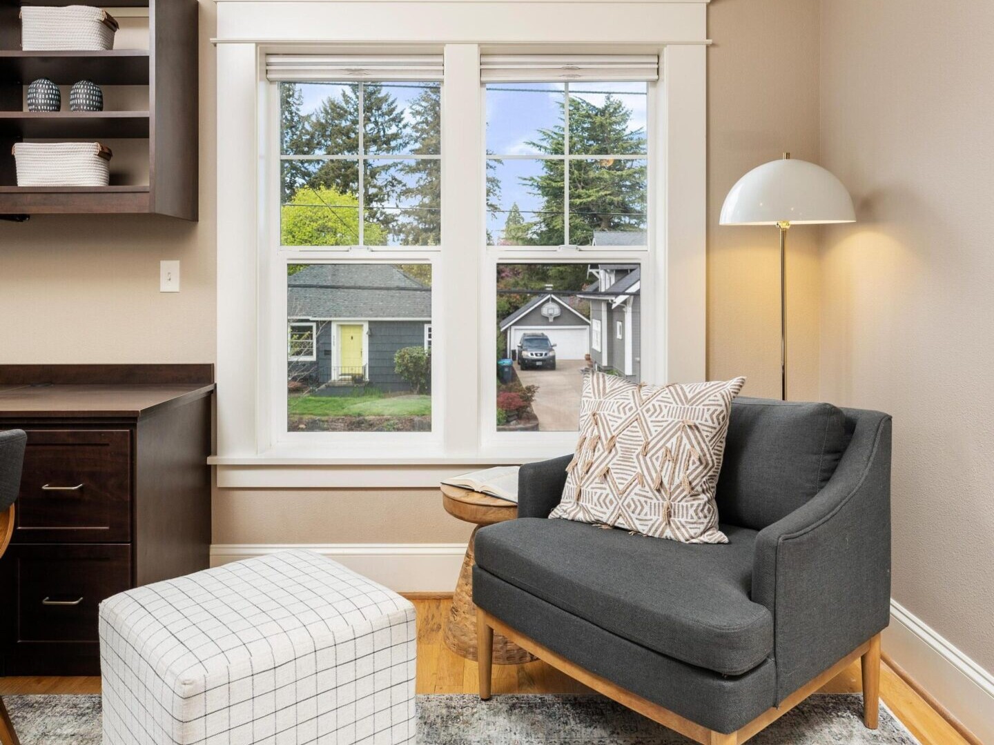 A cozy room in Portland Oregon real estate features a dark gray armchair with a patterned cushion, a plaid ottoman, a floor lamp, and a window overlooking a suburban street. The room has beige walls, wooden flooring, a patterned carpet, and built-in shelving.