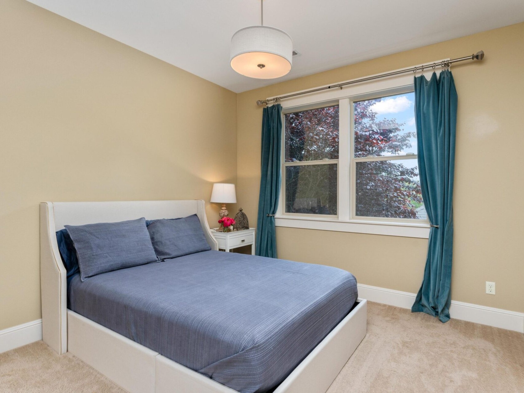 A cozy Portland bedroom with beige walls features a neatly made bed adorned with blue linens. A bedside lamp rests on a white nightstand, and teal curtains frame a window overlooking the trees. Perfect for showcasing Portland Oregon real estate charm, it boasts a chic ceiling light fixture above.
