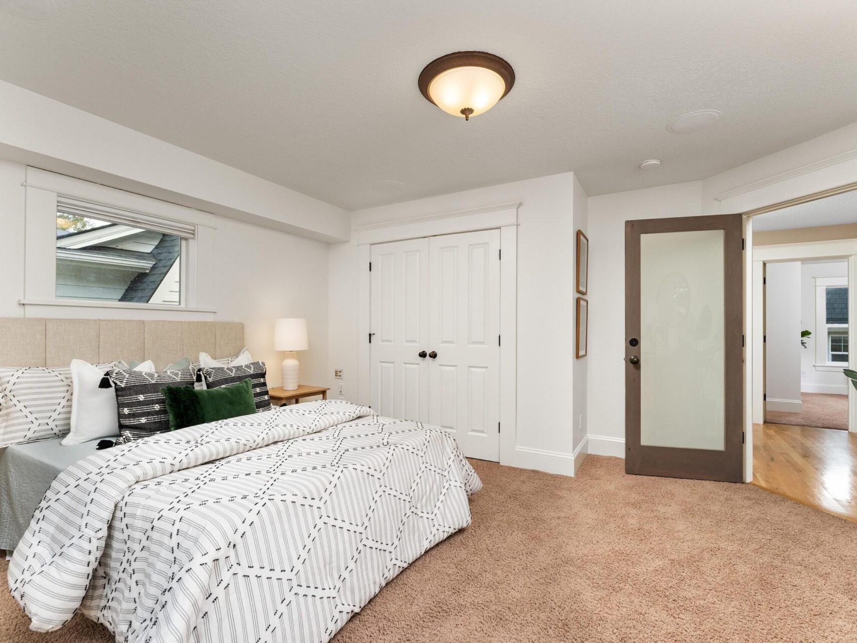 A cozy bedroom in a Portland Oregon Real Estate listing features a large bed with patterned white and gray bedding, green accent pillows, and a bedside lamp. The space includes beige carpet, a closet with double doors, and a frosted glass door leading to the hallway—ideal for any discerning Portland realtor.