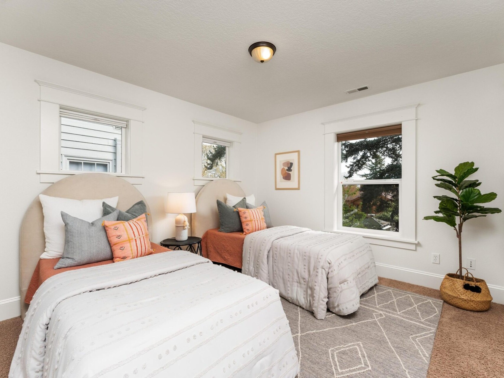 A cozy bedroom with two twin beds, each with white bedding and decorative pillows, is perfect for a Portland Oregon real estate gem. A lamp between the beds sits on a small nightstand. The room features two windows, a leafy plant, and a framed picture above a geometric patterned rug.