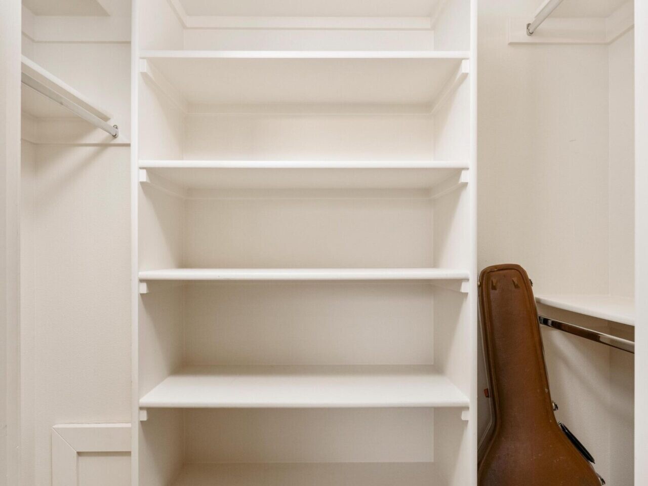 A white closet with empty shelves and a brown guitar case leaning against the wall, emblematic of a Portland Realtor's touch. It features beige carpet flooring and hanging rods on both sides, reflecting the charm of Portland Oregon Real Estate with its warm and inviting design.