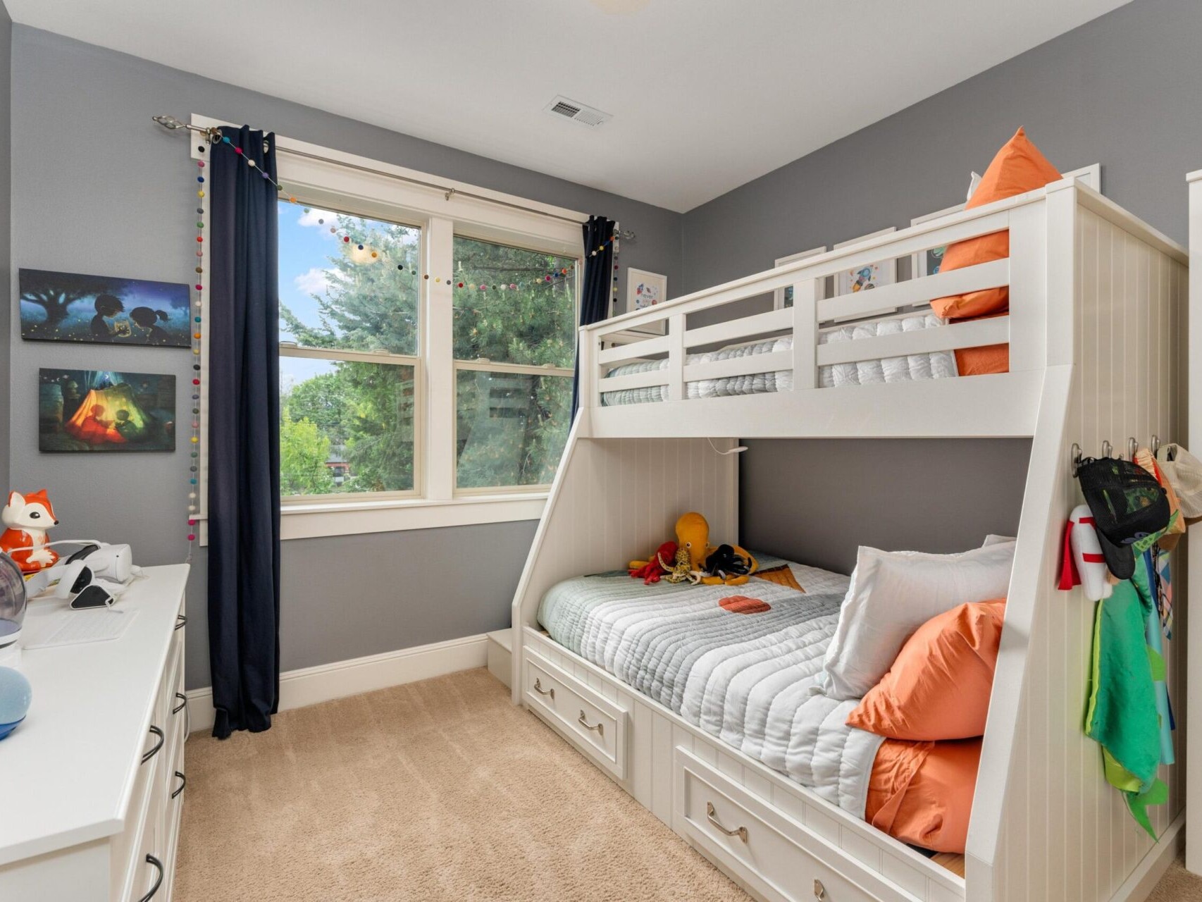 A cozy child's bedroom with gray walls in a charming Portland home features a white bunk bed. The lower bunk boasts orange pillows and a dinosaur stuffed toy. A window with curtains reveals greenery outside, while the room also includes a dresser and small toys on shelves—perfect for any Portland Realtor to showcase!