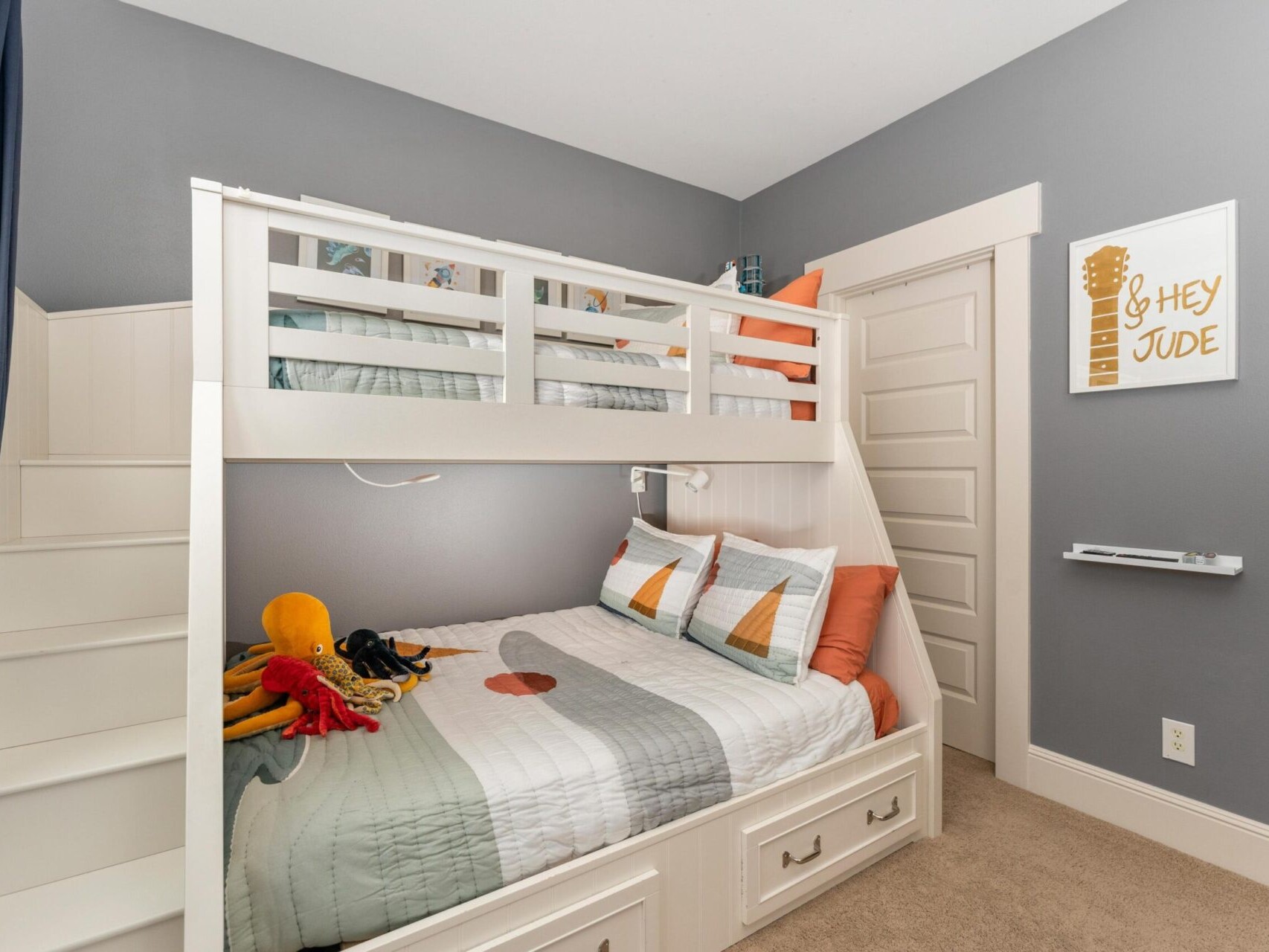 A cozy children's bedroom in a Portland realtor's listing features gray walls and a white bunk bed with stairs. The bedding is adorned with geometric patterns in orange and gray, and a wall sign reads 
