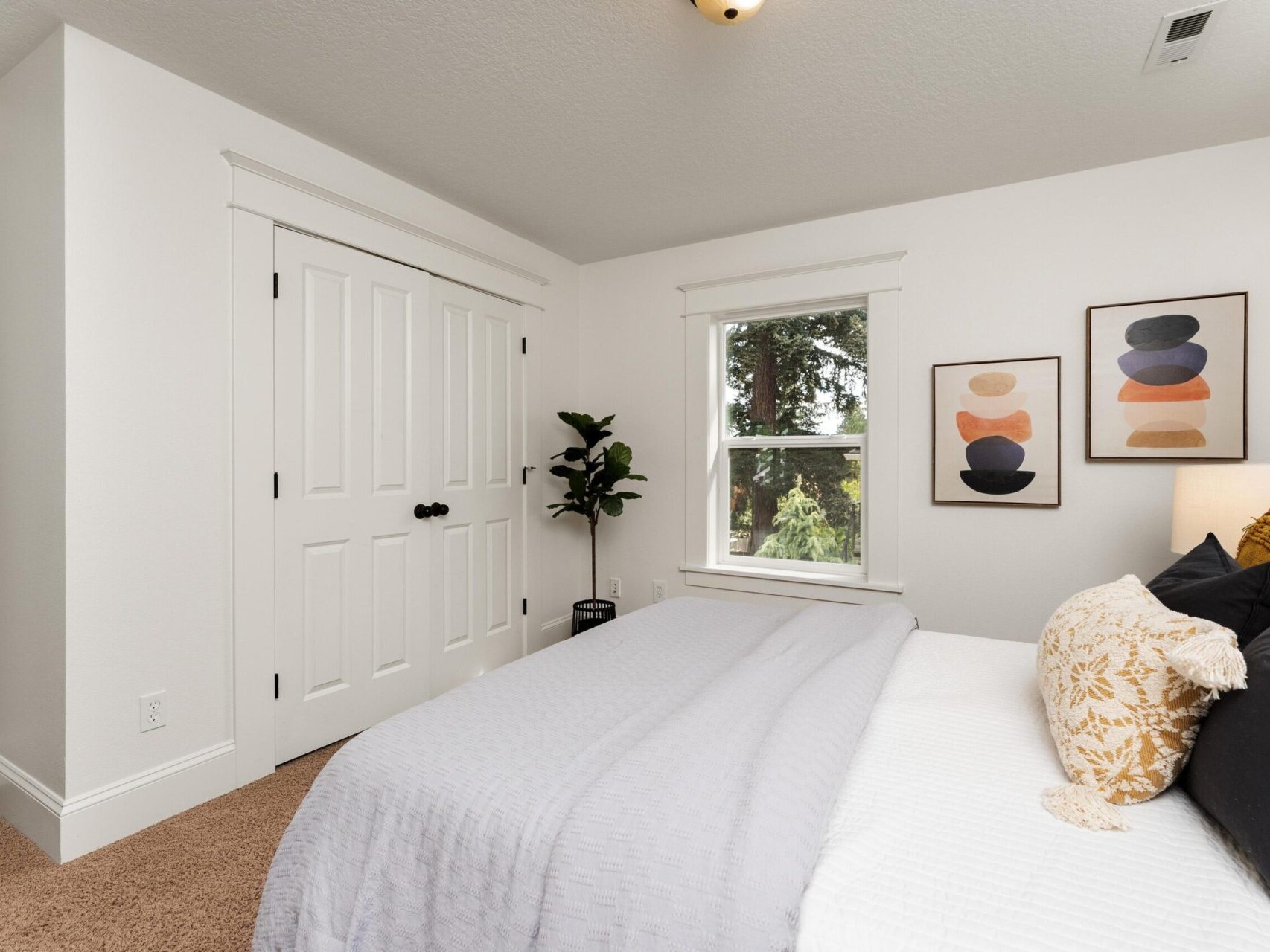 A bright, minimalist bedroom in a stunning Portland Oregon real estate listing features a neatly made bed with white and gray bedding and patterned pillows. A large window lets in natural light, while abstract paintings adorn the walls. Nestled beside the double-door closet is a lush plant.