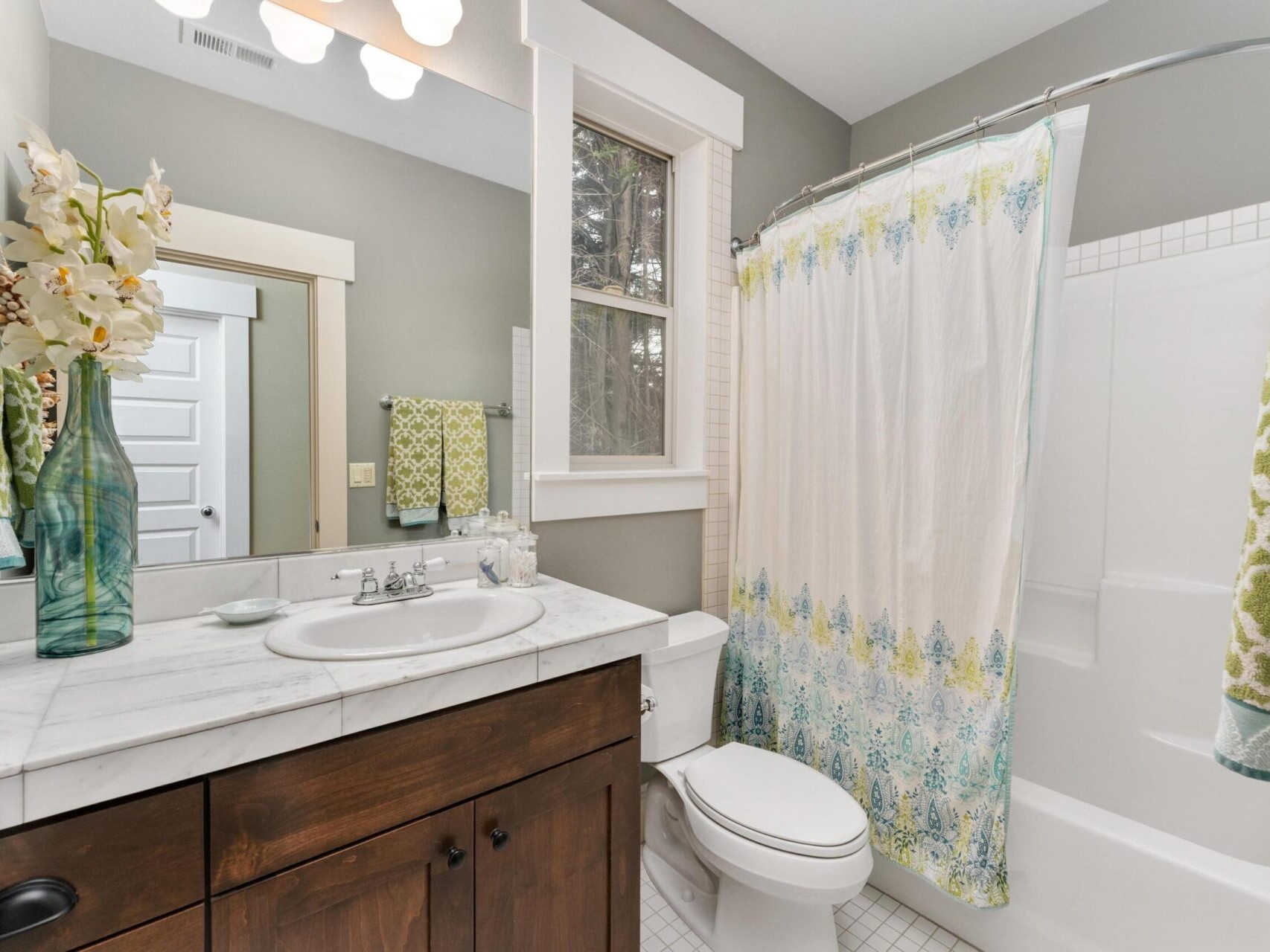 A well-lit bathroom with a wooden vanity and marble countertop exudes Portland Oregon real estate charm. It features a sink, decorative vases, a mirror, a toilet, and a bathtub with a curved shower rod and patterned curtain. Towels hang beside the bathtub, while the window enhances natural light.