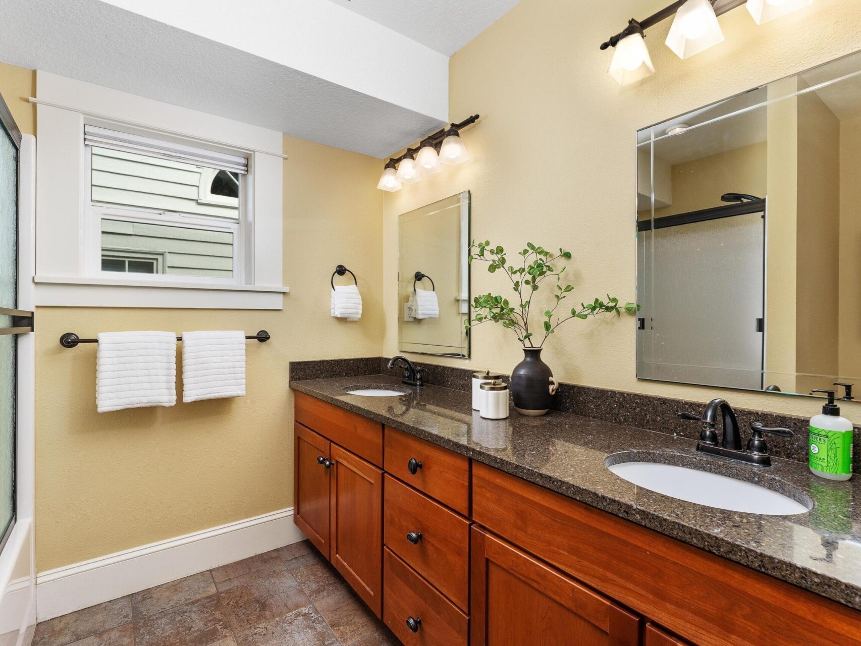 A bathroom with dual sinks and a dark countertop, perfect for Portland Oregon real estate. Wooden cabinets below the sinks and a large mirror reflect under modern fixtures. A black vase with greenery sits between the sinks, while towels hang under a small window on the left.