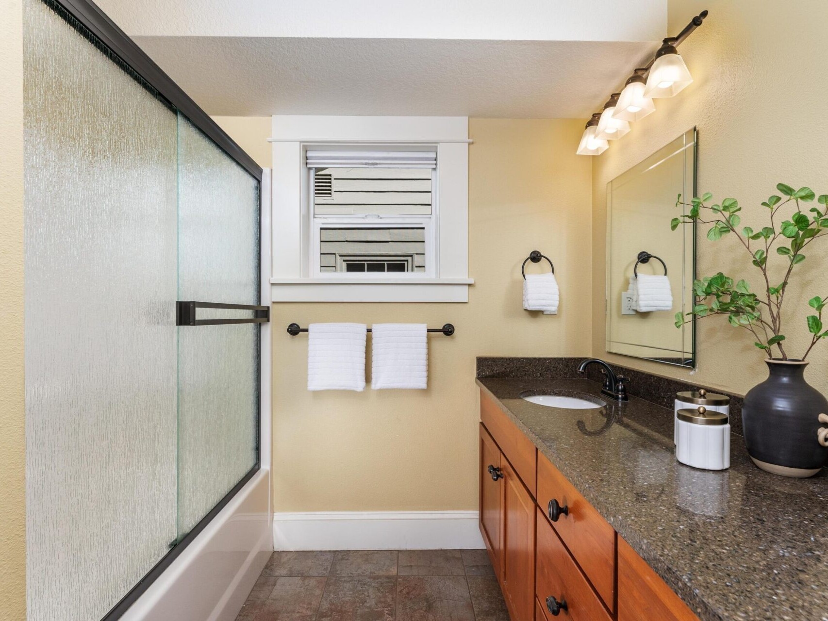 A modern bathroom designed for elegance, featuring a glass-enclosed bathtub on the left and a brown granite countertop with sink. A large wall mirror reflects the sophistication favored by discerning clients in Portland Oregon Real Estate circles. Beige walls complement potted plants and dual towel racks under warm overhead lighting.