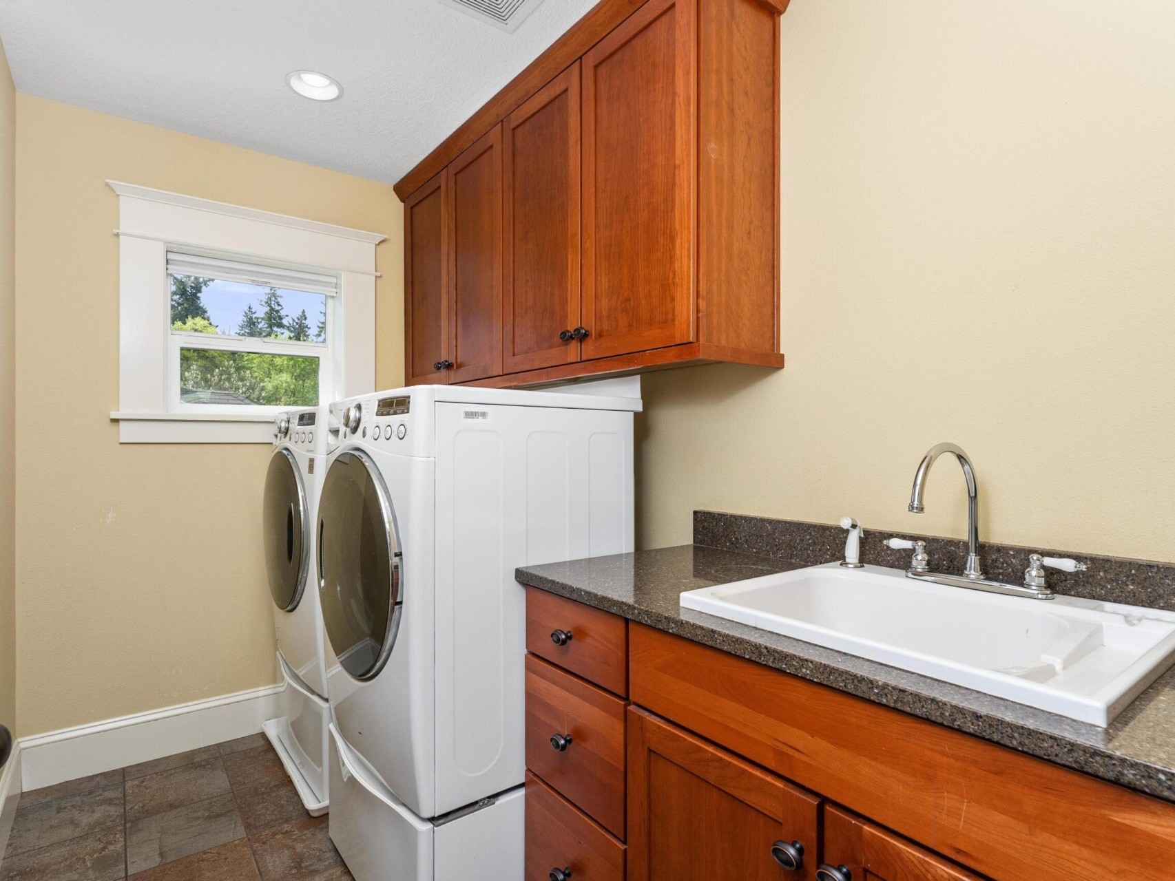 This Portland, Oregon real estate gem features a laundry room with a front-loading washer and dryer on the left beneath a brown cabinet. On the right, find a counter with a sink. Light streams in through a small window set against the cream-colored wall above. Ideal for any discerning buyer!
