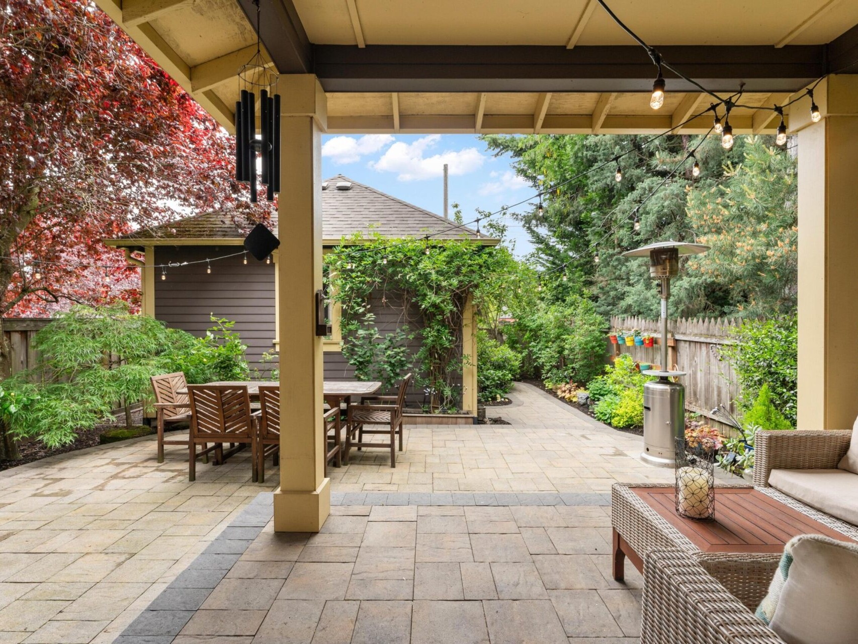 A cozy backyard patio in Portland, Oregon real estate features cushioned seating, a wooden dining table, and string lights overhead. The space is surrounded by lush greenery, with a red-leafed tree and a small garden shed in the background.