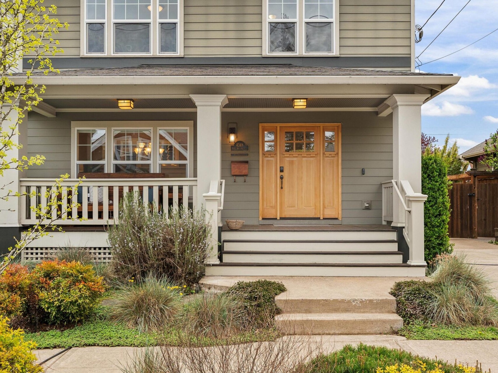 Front view of a two-story Portland home with a gray exterior and wooden door. The porch, adorned with white railings and two overhead lights, invites you in. The yard features green shrubs and a small tree, with a concrete pathway leading to the entrance—a true gem in Portland Oregon real estate.