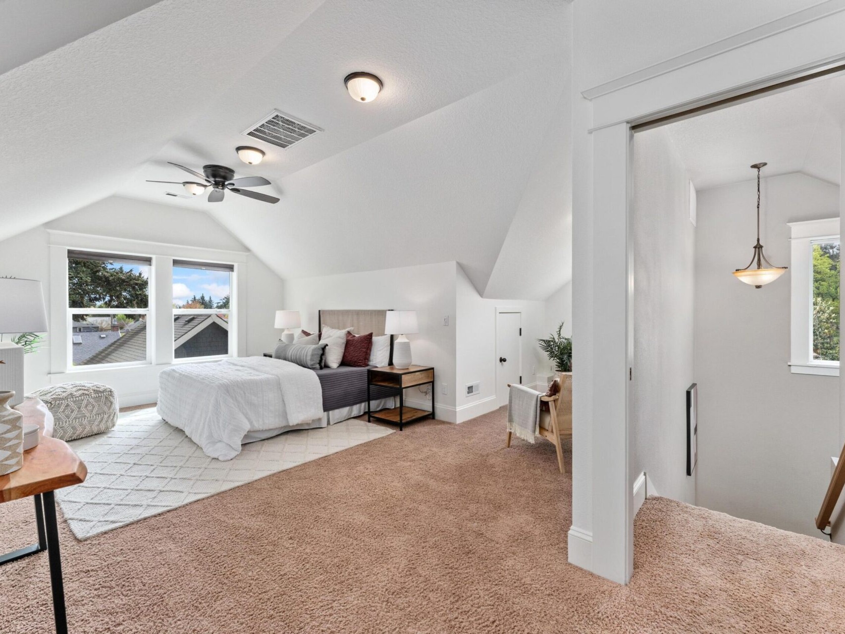 A bright attic bedroom in this charming Portland Oregon real estate listing features sloped ceilings, a cozy bed, and a white area rug on beige carpet. A ceiling fan hangs above the sitting area near the staircase. Large windows flood the space with natural light and offer a view of the trees.