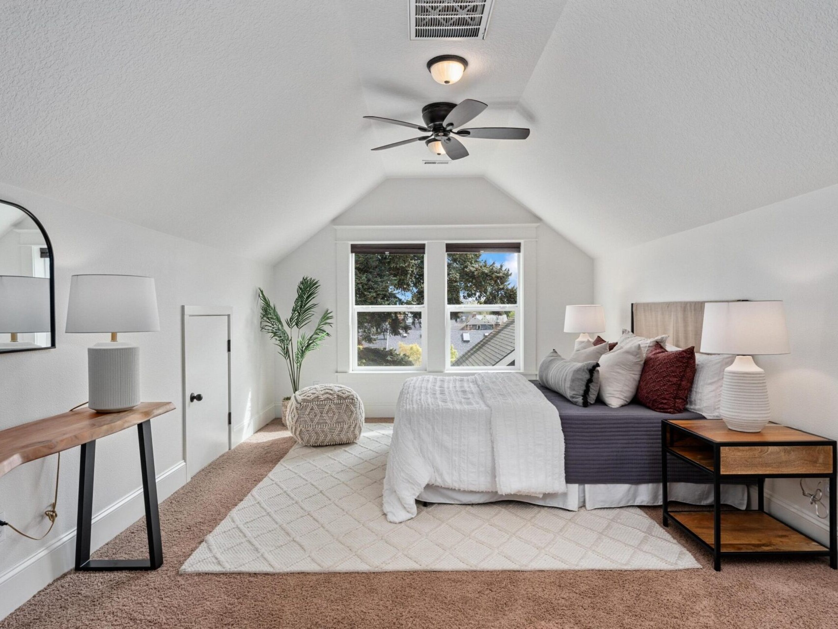 A cozy attic bedroom in Portland Oregon real estate boasts a sloped ceiling, featuring a bed with white and gray bedding, a wooden nightstand with lamps, a large mirror on a console table, a pouf, and a plant. A ceiling fan hangs above, and a window offers outdoor views.